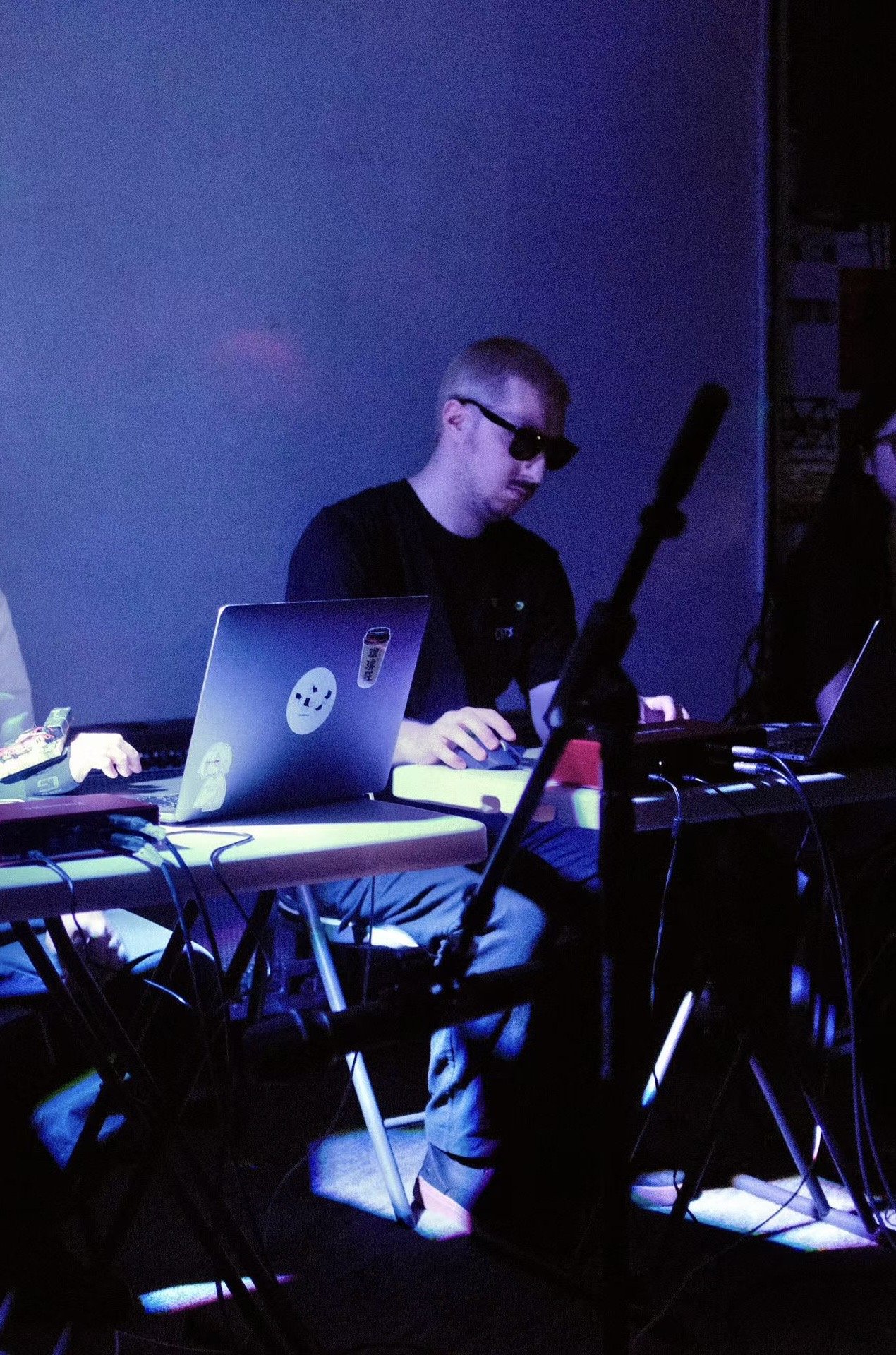 A person wearing sunglasses working on a laptop at a table with multiple electronic devices and cables, in a dimly lit room with blue and purple lighting.