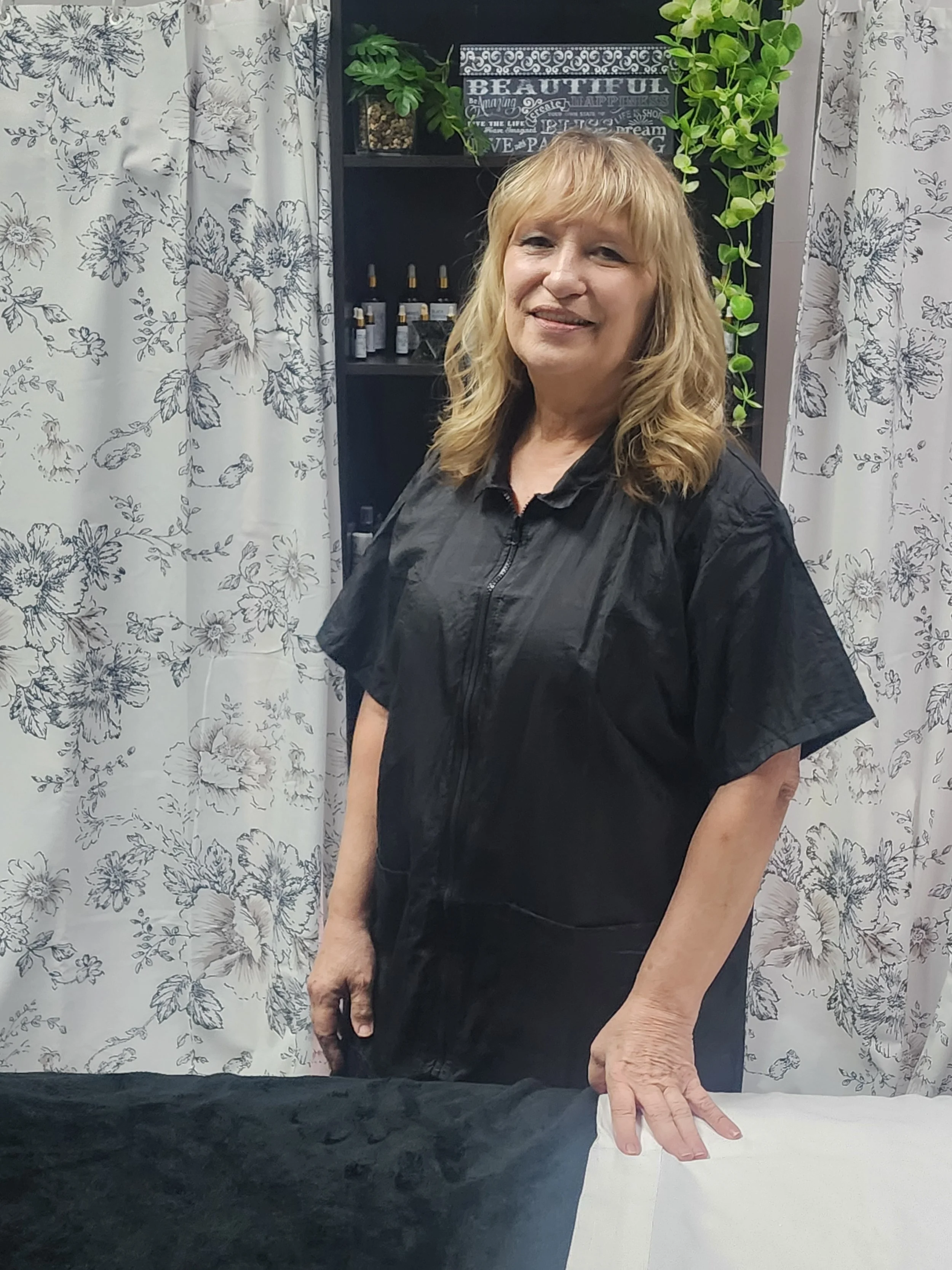 A middle-aged woman with blonde hair styled in loose waves, smiling, stands in front of a decorative black shelf with green plants and a sign that reads "BEAUTIFUL". She is wearing a black shirt and is touching a table covered with white and black fabric. The background features white curtains with black floral patterns.