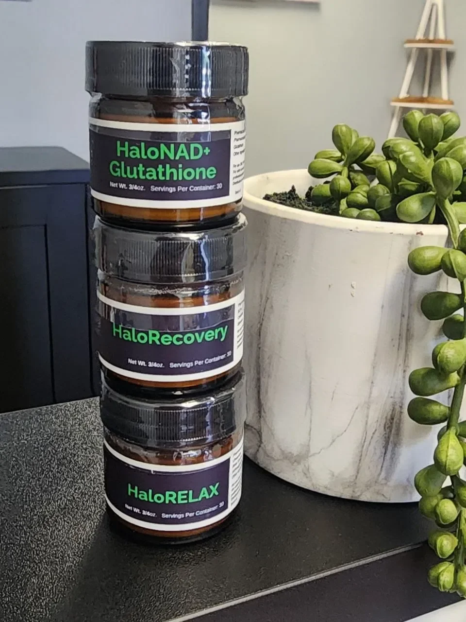 Three bottles labeled 'HaloNAD+ Glutathione,' 'HaloRecovery,' and 'HaloRELAX' stacked next to a potted succulent plant.