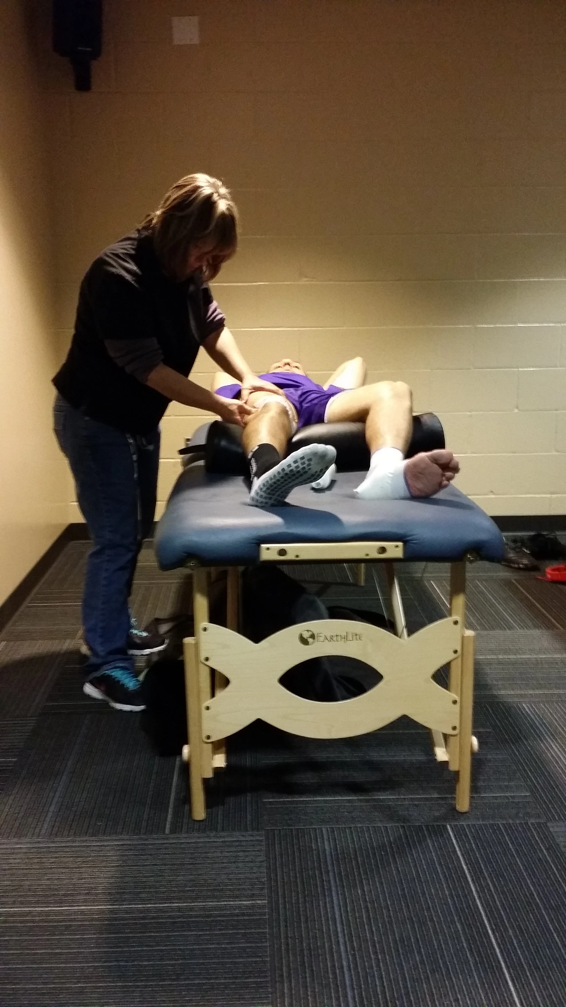 Client lying on a massage table receiving medical treatment as Emma Anderson tends to their leg, in an indoor setting.