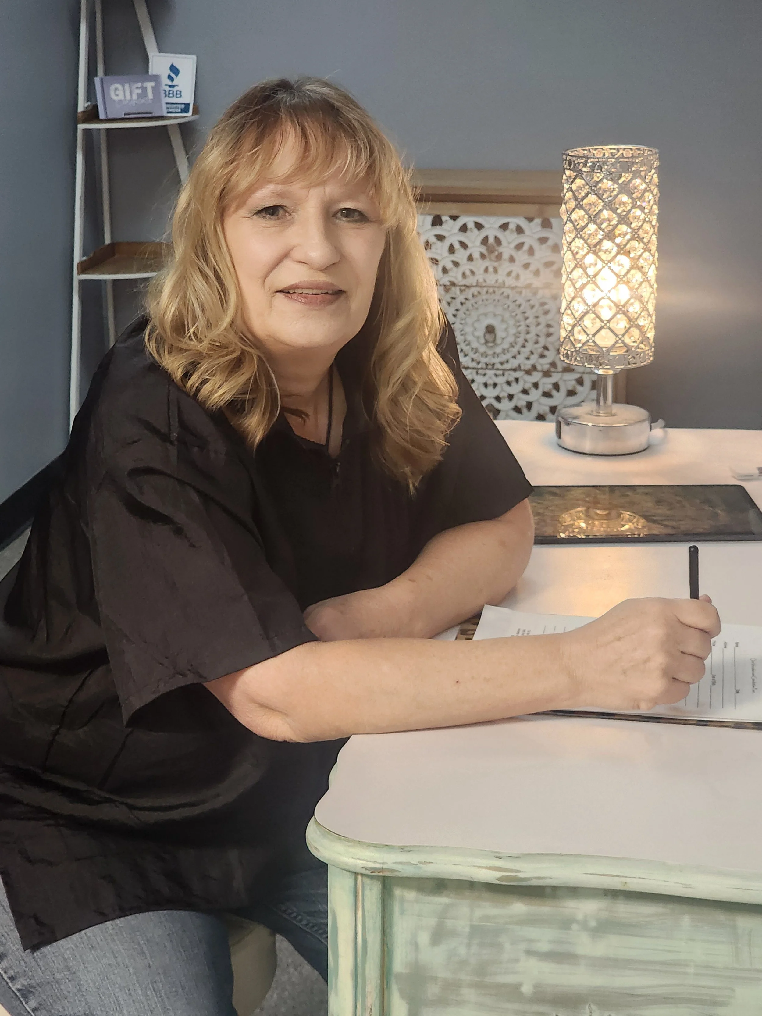 Emma Anderson sits at a table writing on a paper with a pen. There is a decorative lamp with a woven pattern behind her, emitting warm light, and a piece of art or decorative panel with a circular pattern on the wall.