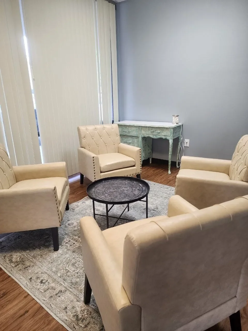 A cozy sitting area with five beige upholstered armchairs arranged in a circle around a small black round coffee table. There is a light-colored rug underneath the table, and in the background, a light green distressed wooden side table is placed against a pale blue wall. Vertical blinds cover the window, allowing some light to filter into the room.