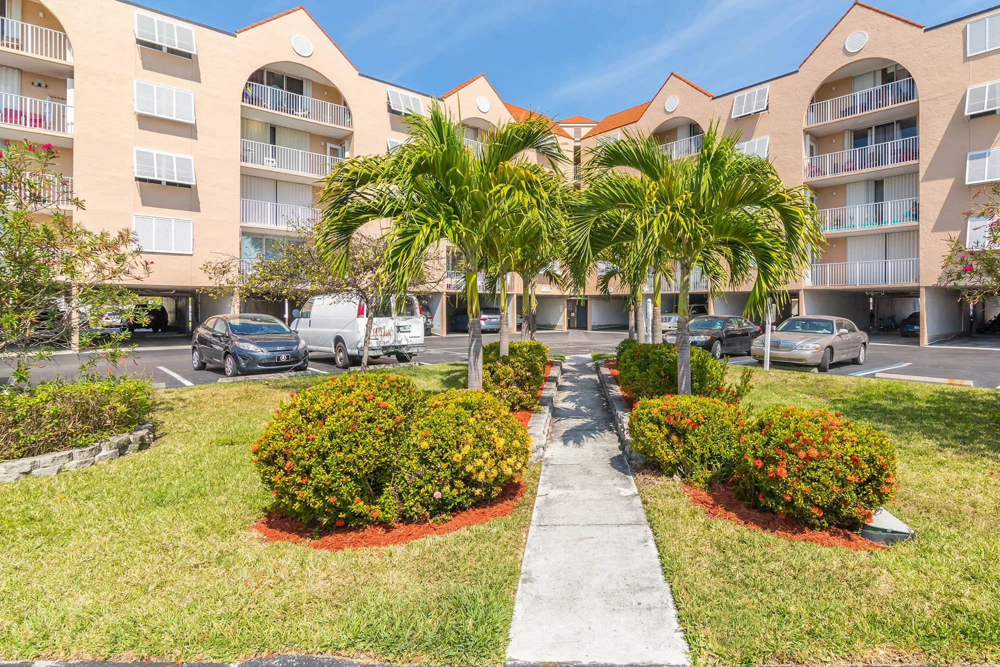 View of a residential apartment complex with a parking lot and landscaped area with palm trees, bushes, and a pathway.