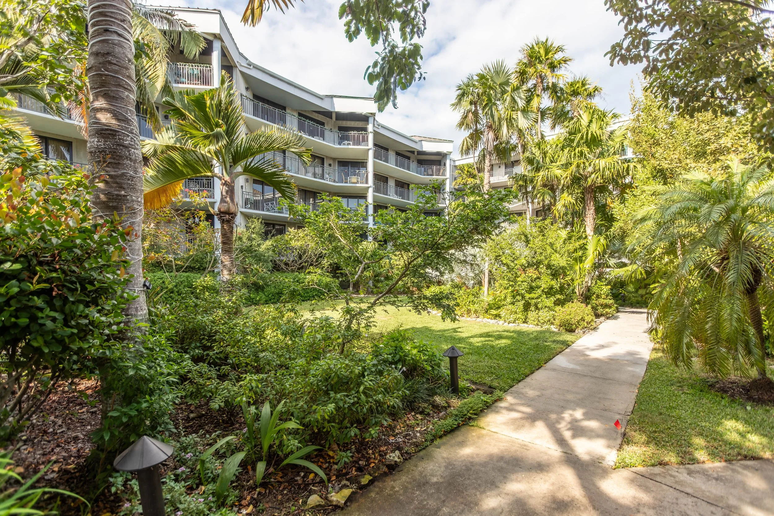 A landscaped garden with green shrubs, trees, and a concrete pathway leading to a multi-story residential building with balconies in the background, surrounded by lush tropical plants and palm trees.
