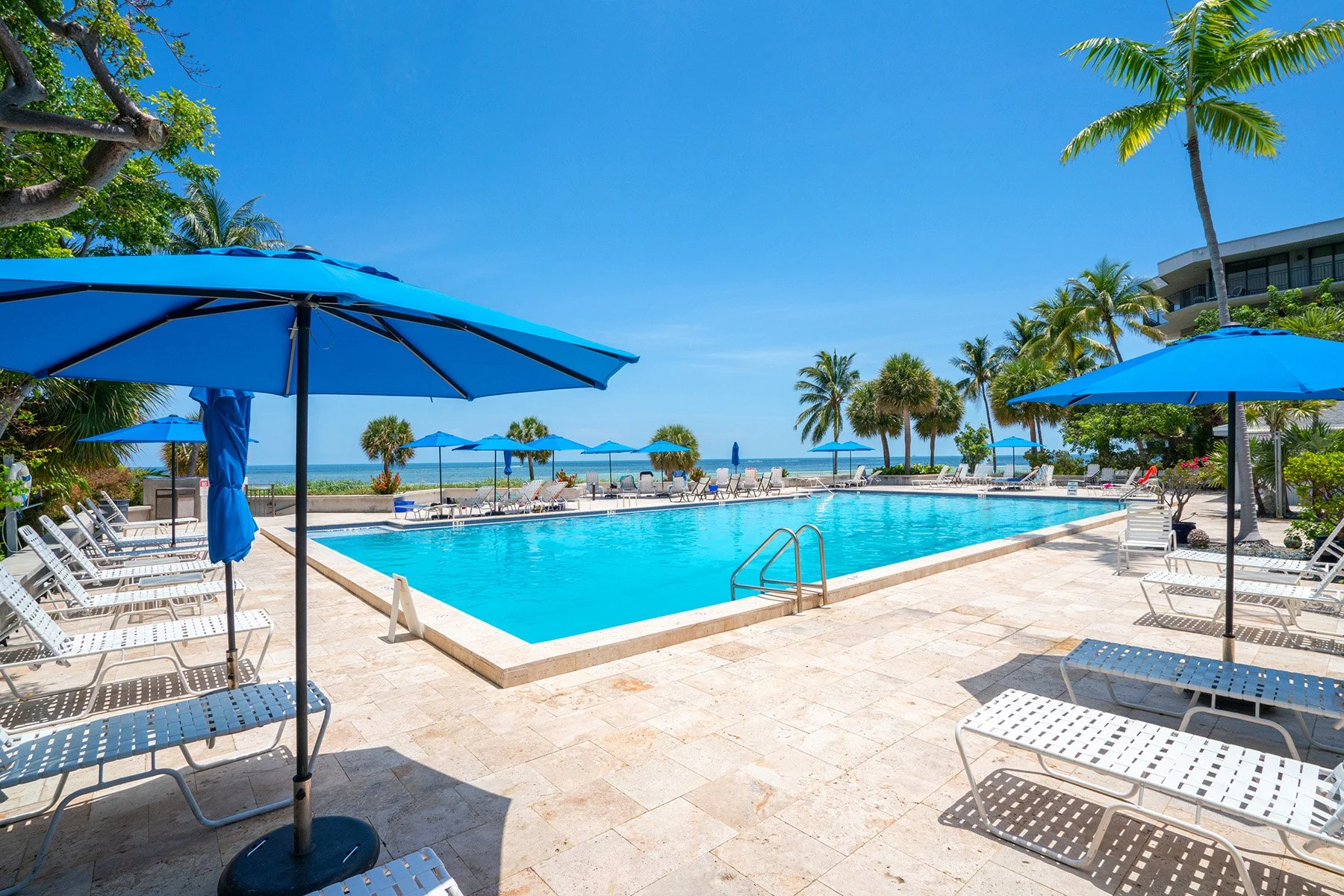 An outdoor swimming pool area at a tropical resort with blue umbrellas, lounge chairs, palm trees, and a view of the beach and ocean in the background during a sunny day.