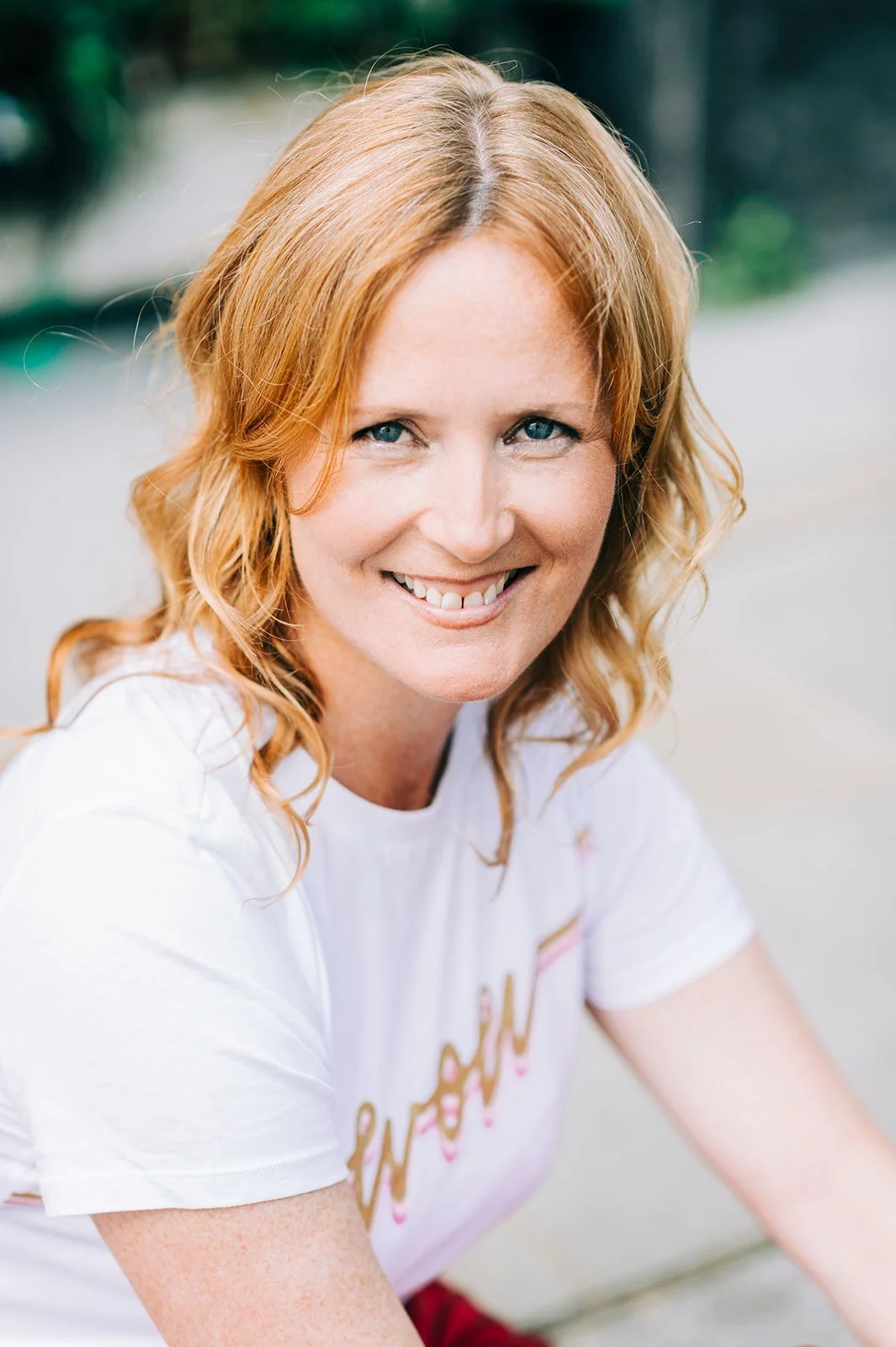 A smiling woman with wavy red hair and blue eyes, wearing a white t-shirt, outdoors in natural light.