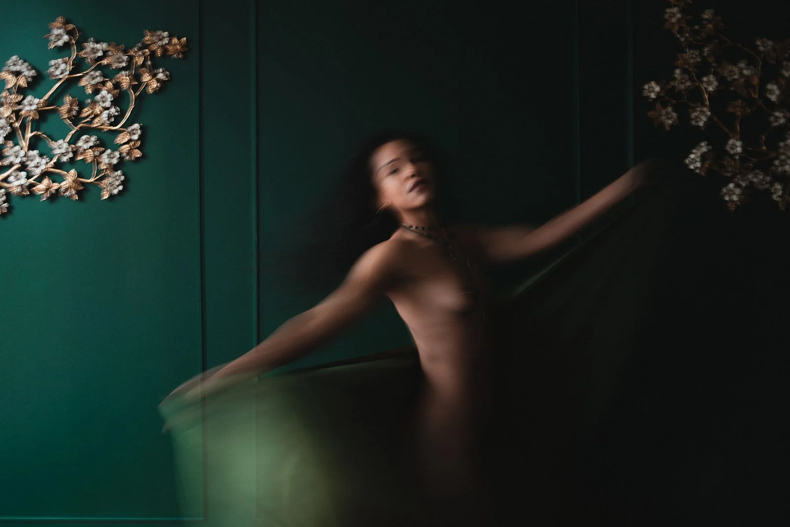 a woman is shown in blurry motion moving from the left of the image to the right, holding a green fabric and spreading her arms wide. She is set on a green background with floral wall decor on either side of the frame.