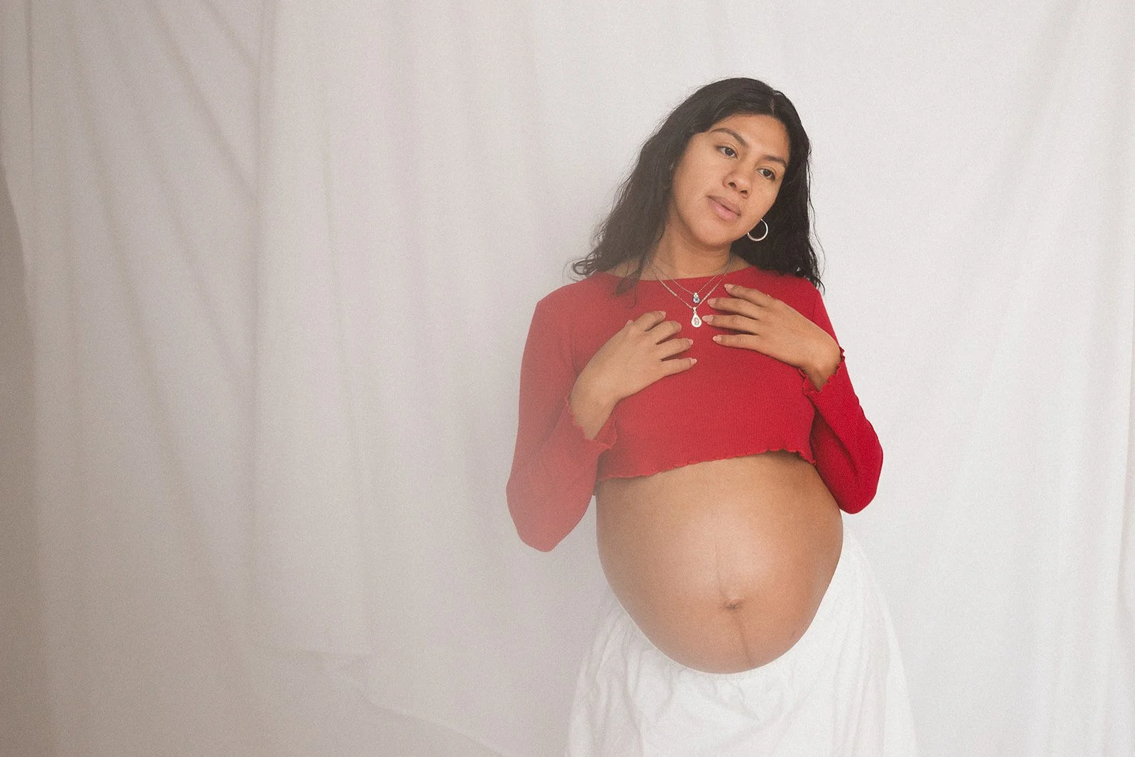 a pregnant latina woman photographed on a white fabric backdrop. She looks off to the right of the viewer, and has her hands at her collarbones.