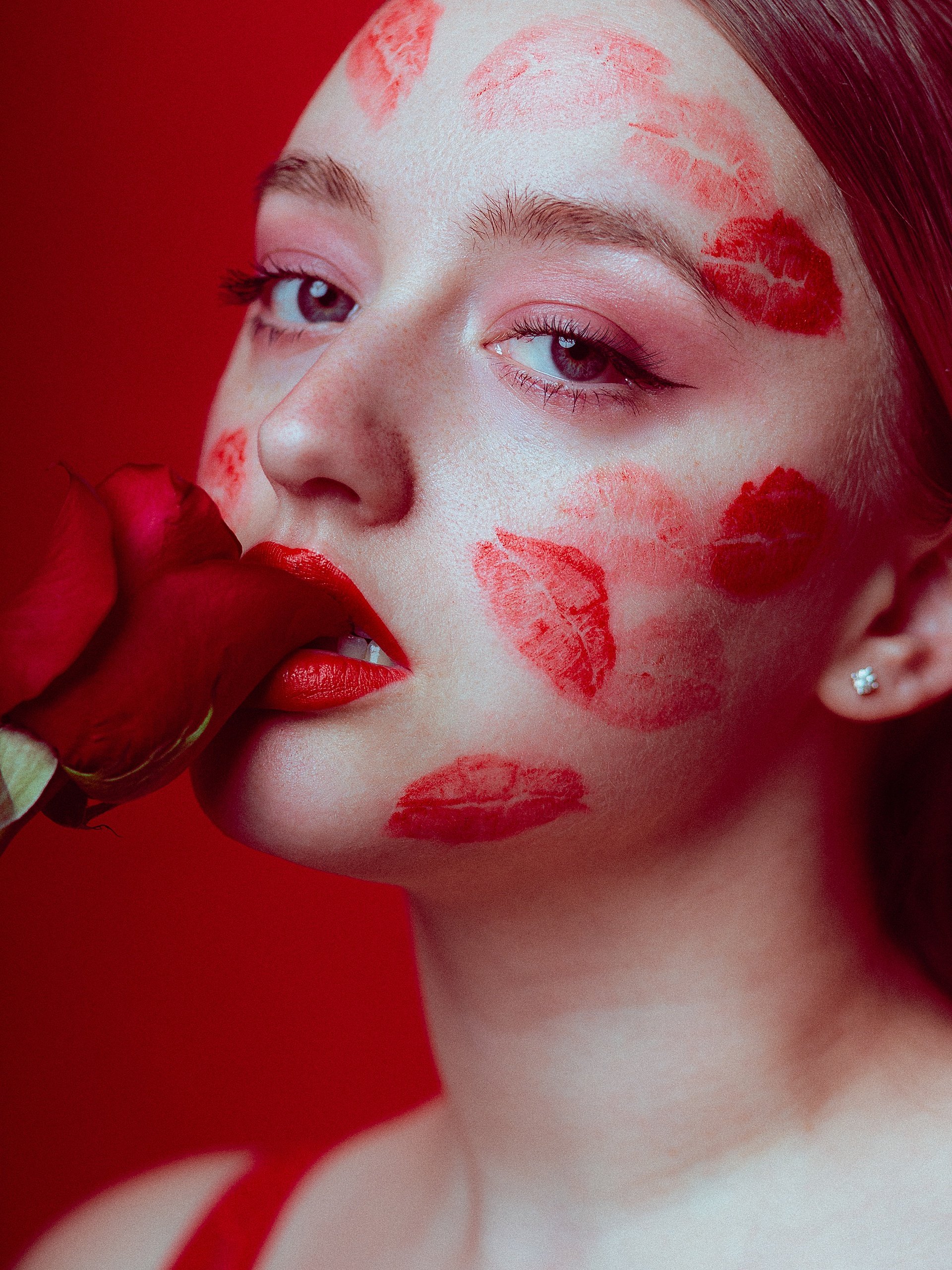 close up photo of a woman biting the petal of a red rose. She has red lip marks all over her face, and she is photographed against a red studio backdrop.