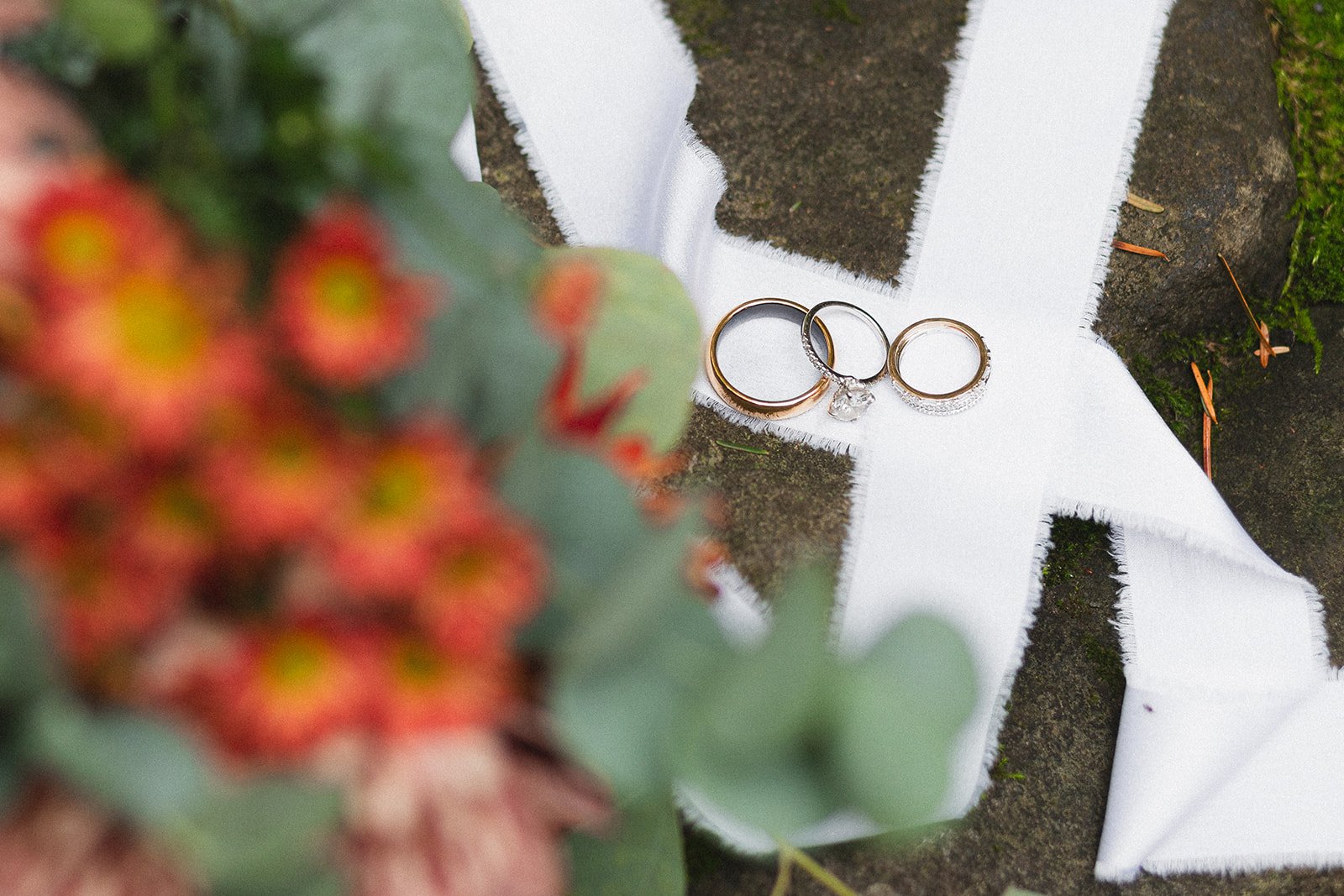 a blurred bouquet of orange flowers takes up the left half of the image. In the right half, 3 wedding rings are laid out on a white ribbon.