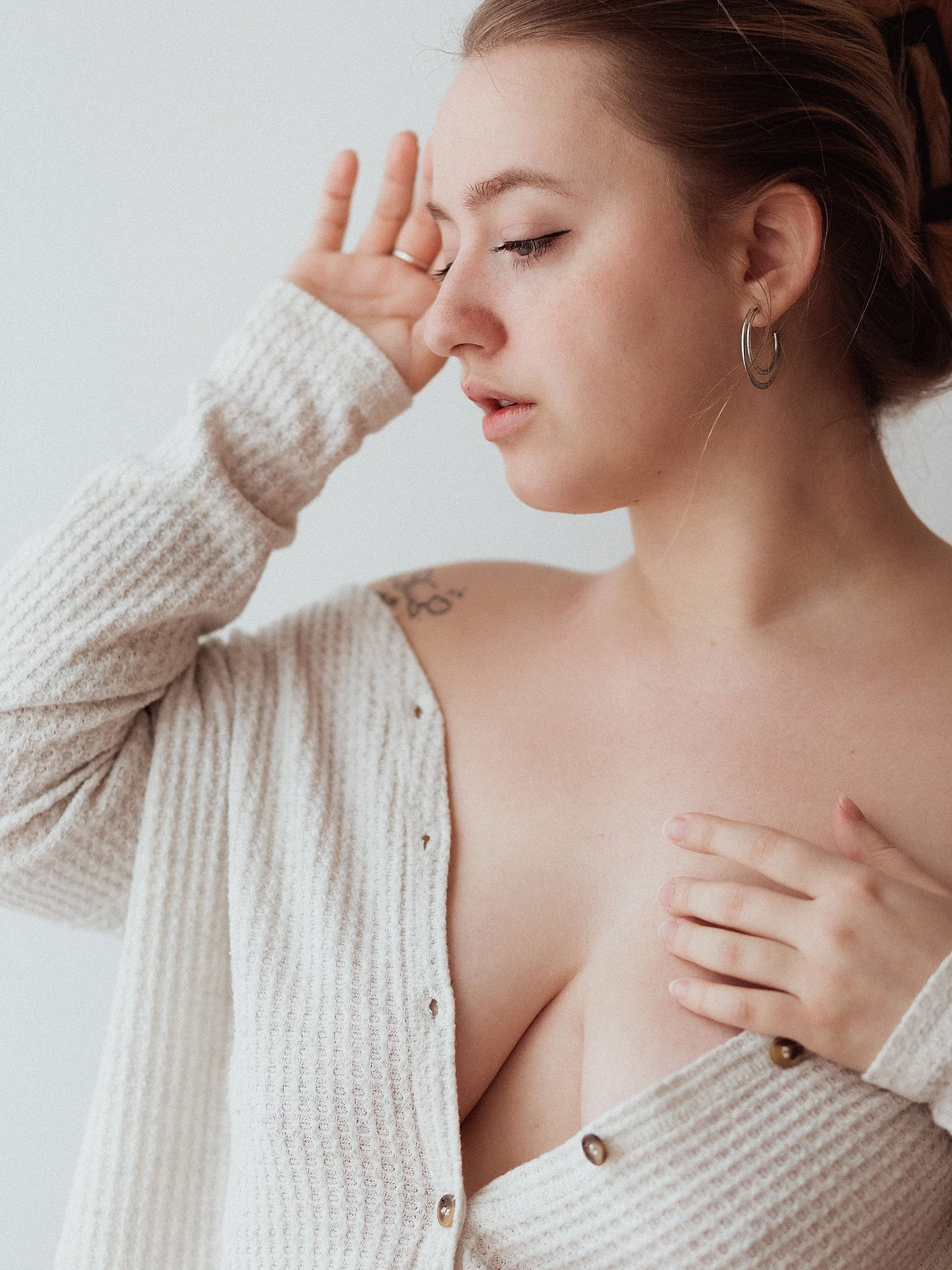 photo of a woman wearing a white sweater partially unbuttoned down the front. She poses for a boudoir self portrait against a plain white wall.