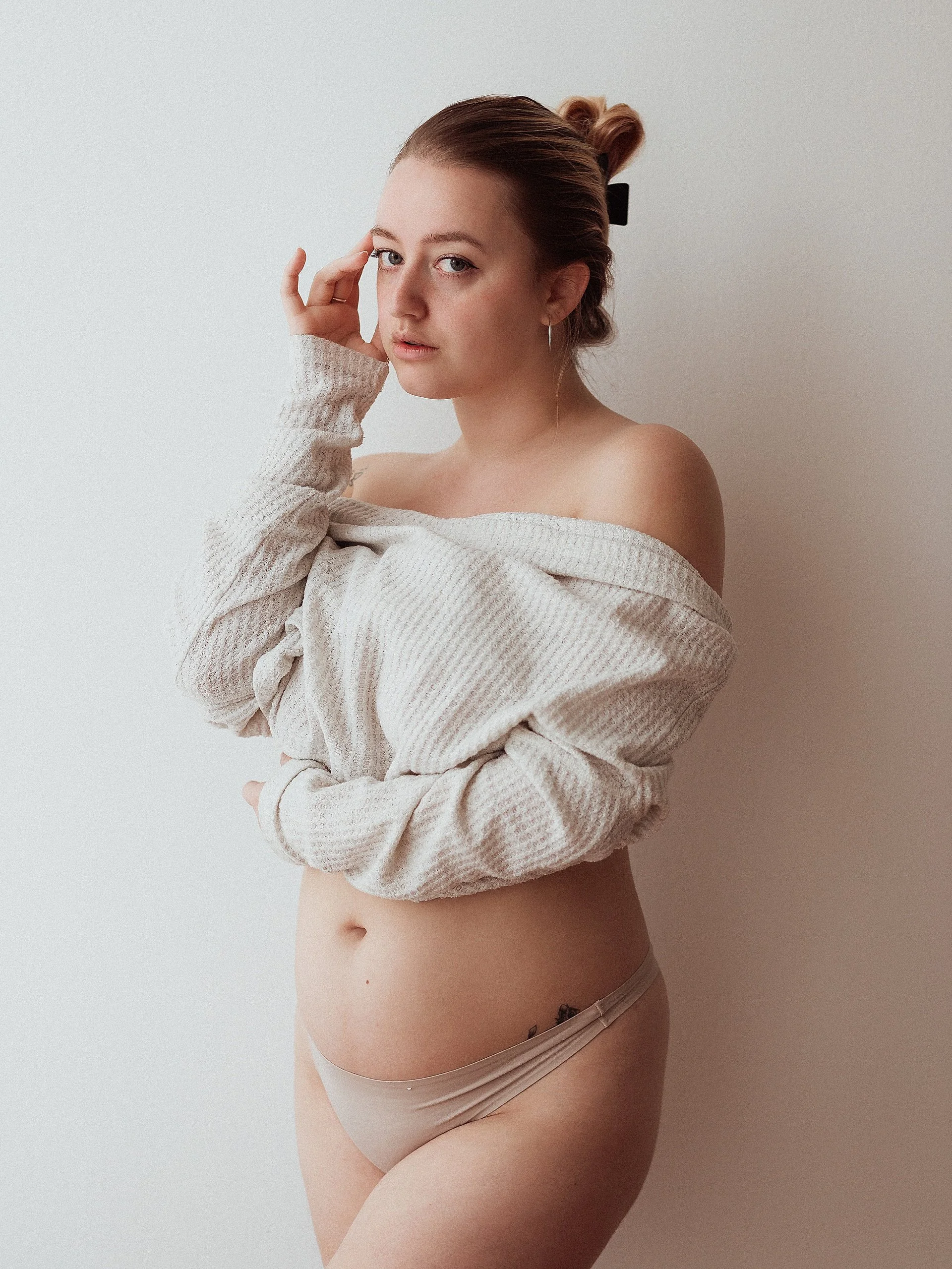 photo of woman standing against a plain white wall. She is posed with her sweater pulled halfway up her arms, leaving her shoulders exposed. She has one hand touching the opposite elbow, and the other rested on her temple. Her hair is up in a clip.