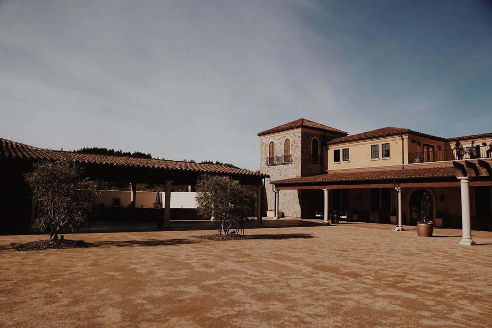 A spacious courtyard with a tan dirt ground, two small trees, and a large building with a tiled roof, arches, and a tower. The sky is mostly cloudy.