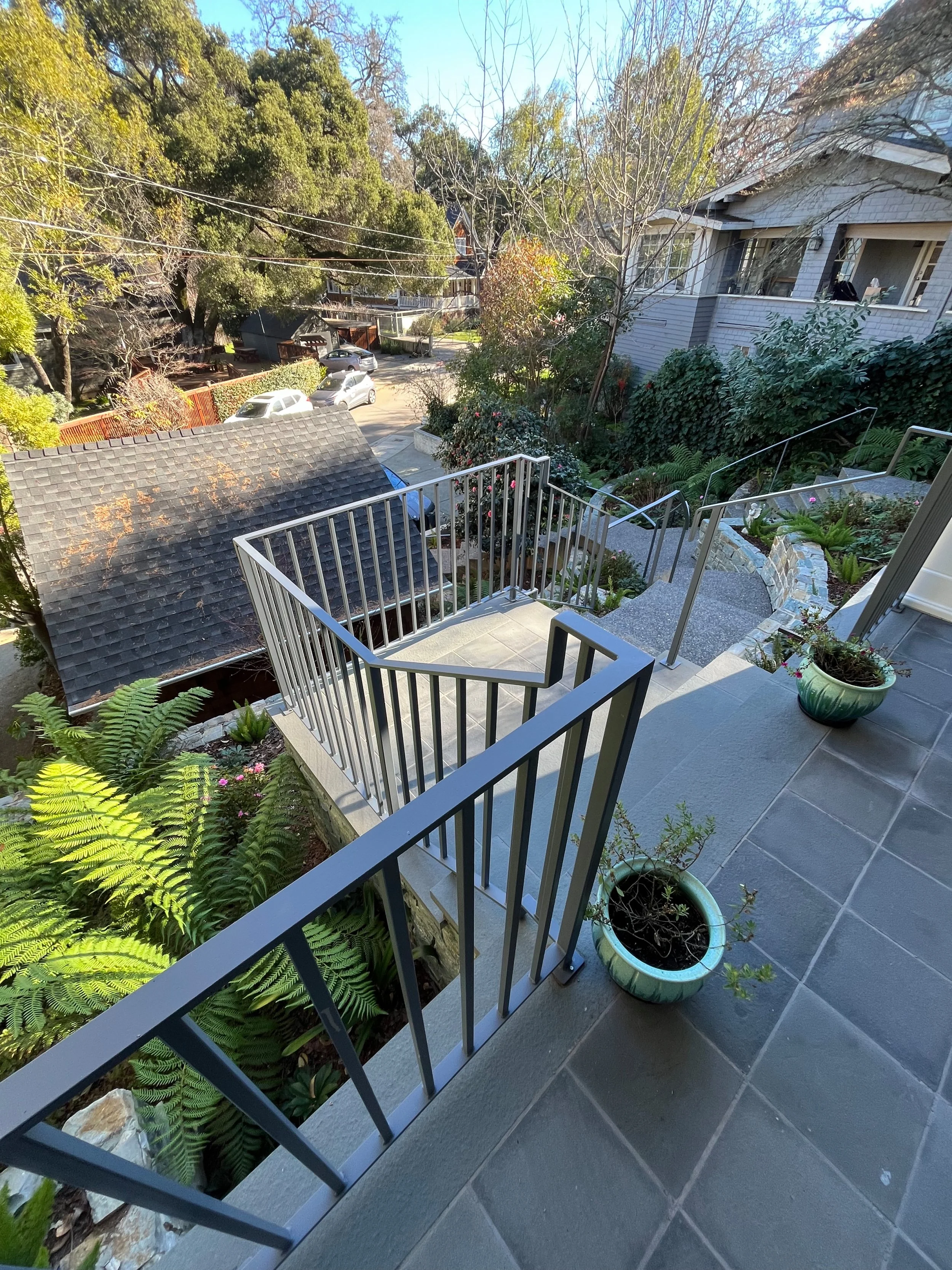 View from a balcony with potted plants and a staircase leading down to a garden with lush greenery and neighboring houses in a suburban neighborhood.