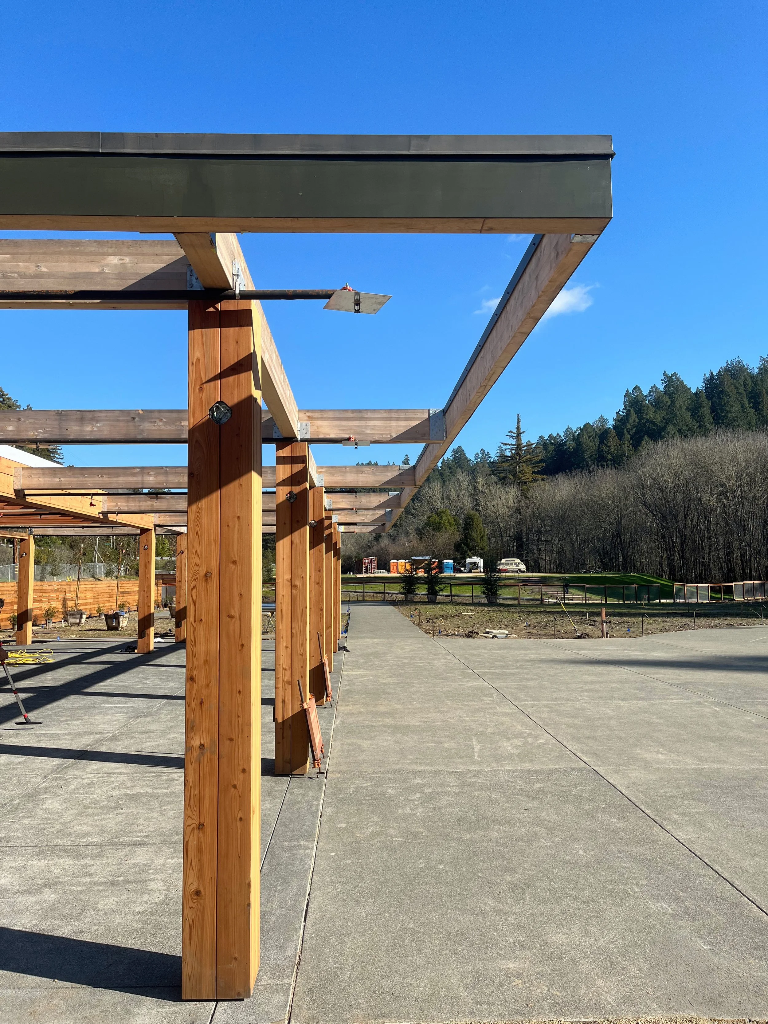 Construction site with wooden framework for a building, clear blue sky, open area with concrete pavement, and trees in the background.
