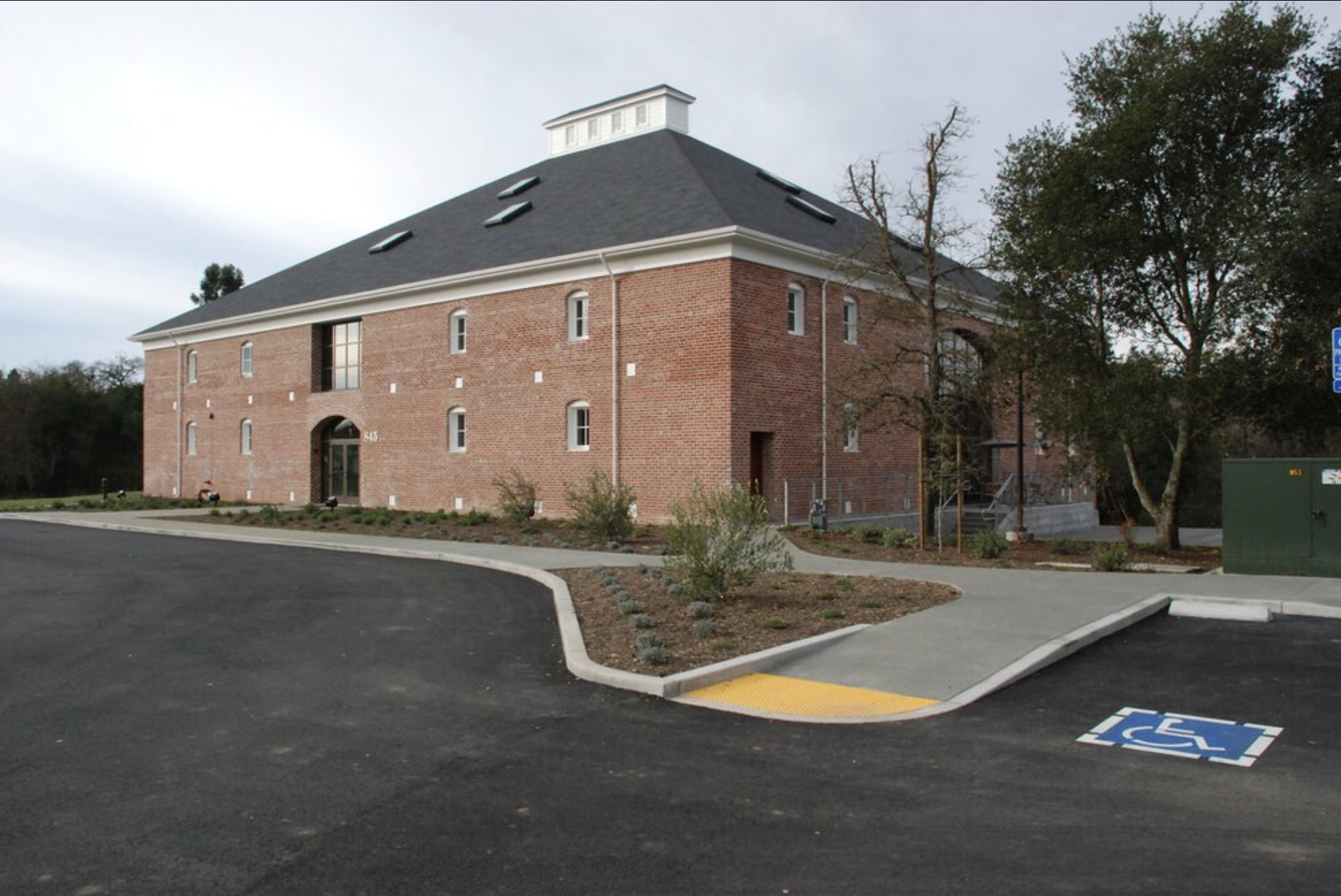 A brick apartment building with a dark gray roof, small windows, and a covered entrance. In front is a paved parking lot with a handicap parking space marked by blue and white paint, and a ramp with a yellow textured surface for accessibility. Trees and landscaping are visible around the building.