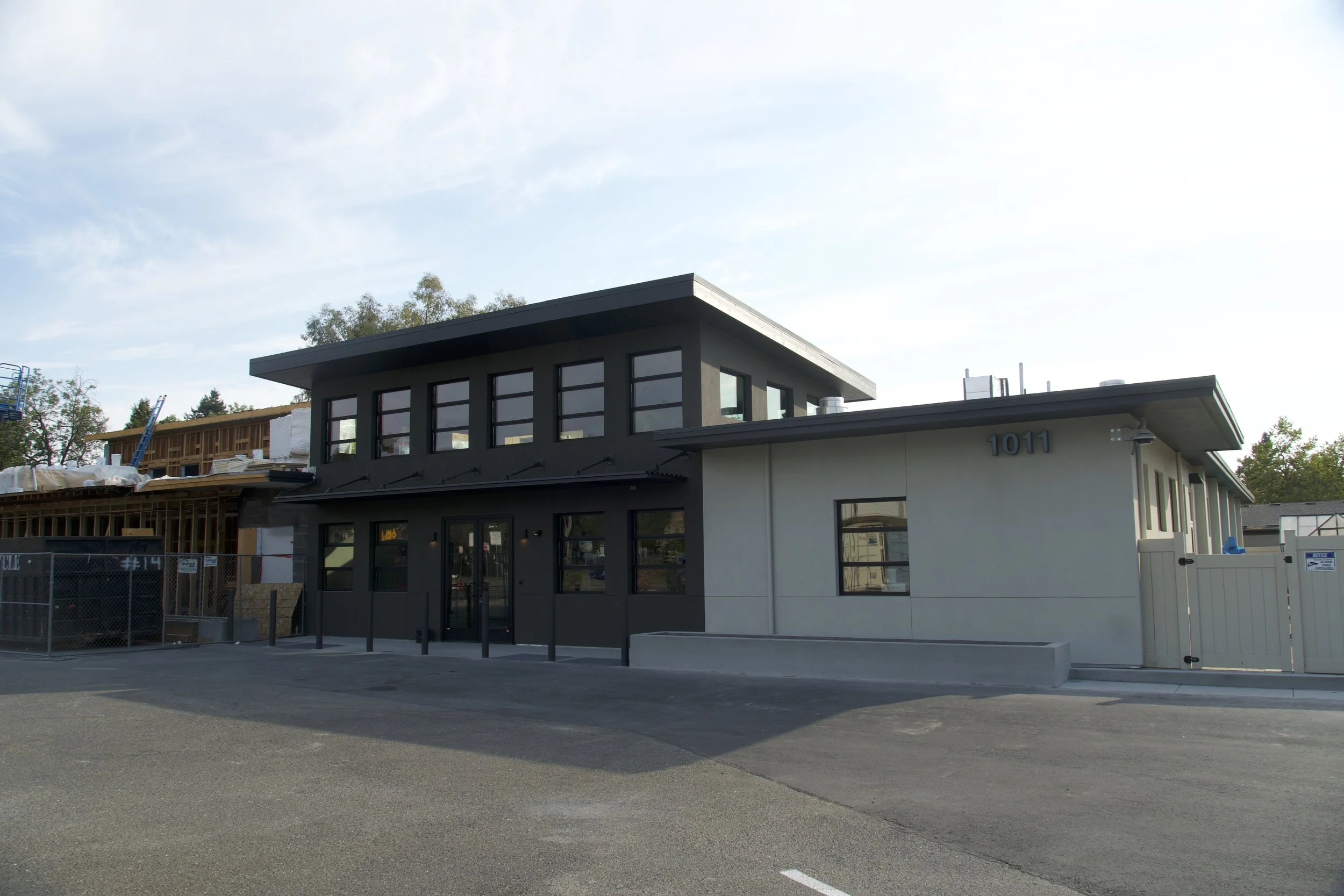 A modern two-story building with gray and white exterior walls, large windows, and a flat roof, located in a parking lot.