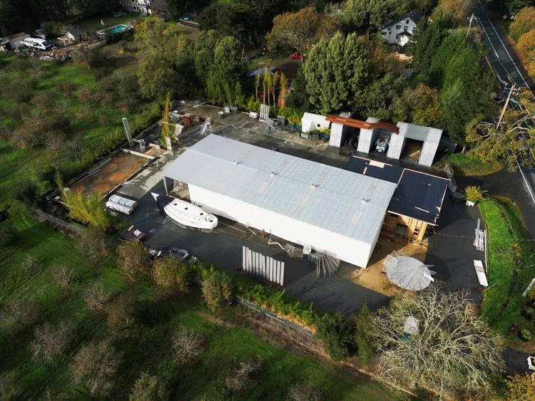 Aerial view of a large white industrial or warehouse building surrounded by greenery, trees, and a few parked boats.