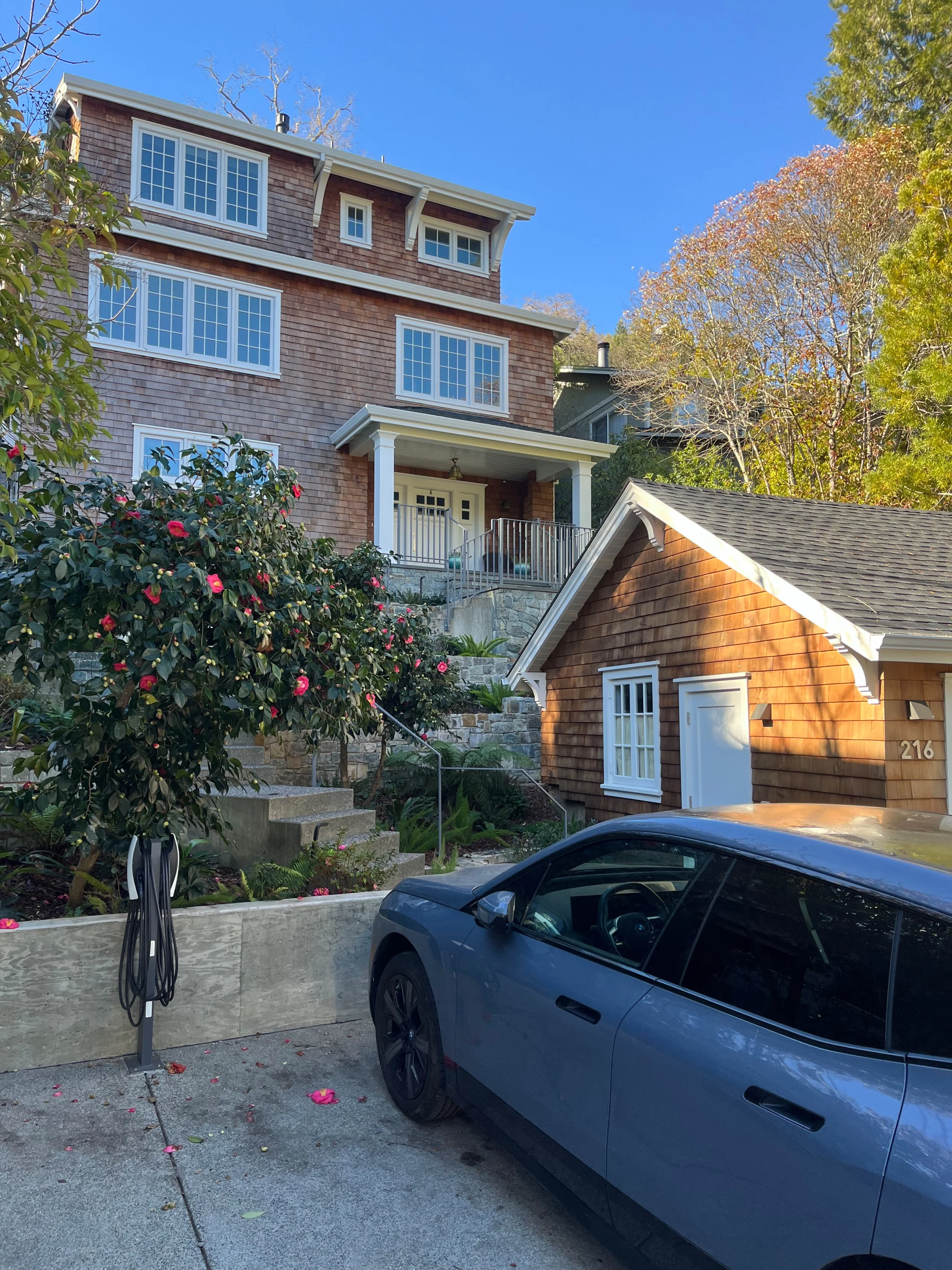 A driveway with a blue car parked in front of a wooden house with shingle siding. A large camellia bush with pink flowers is on the left. There are stairs and a railing leading up to a porch on a larger multi-story house in the background. Trees with autumn foliage are visible.
