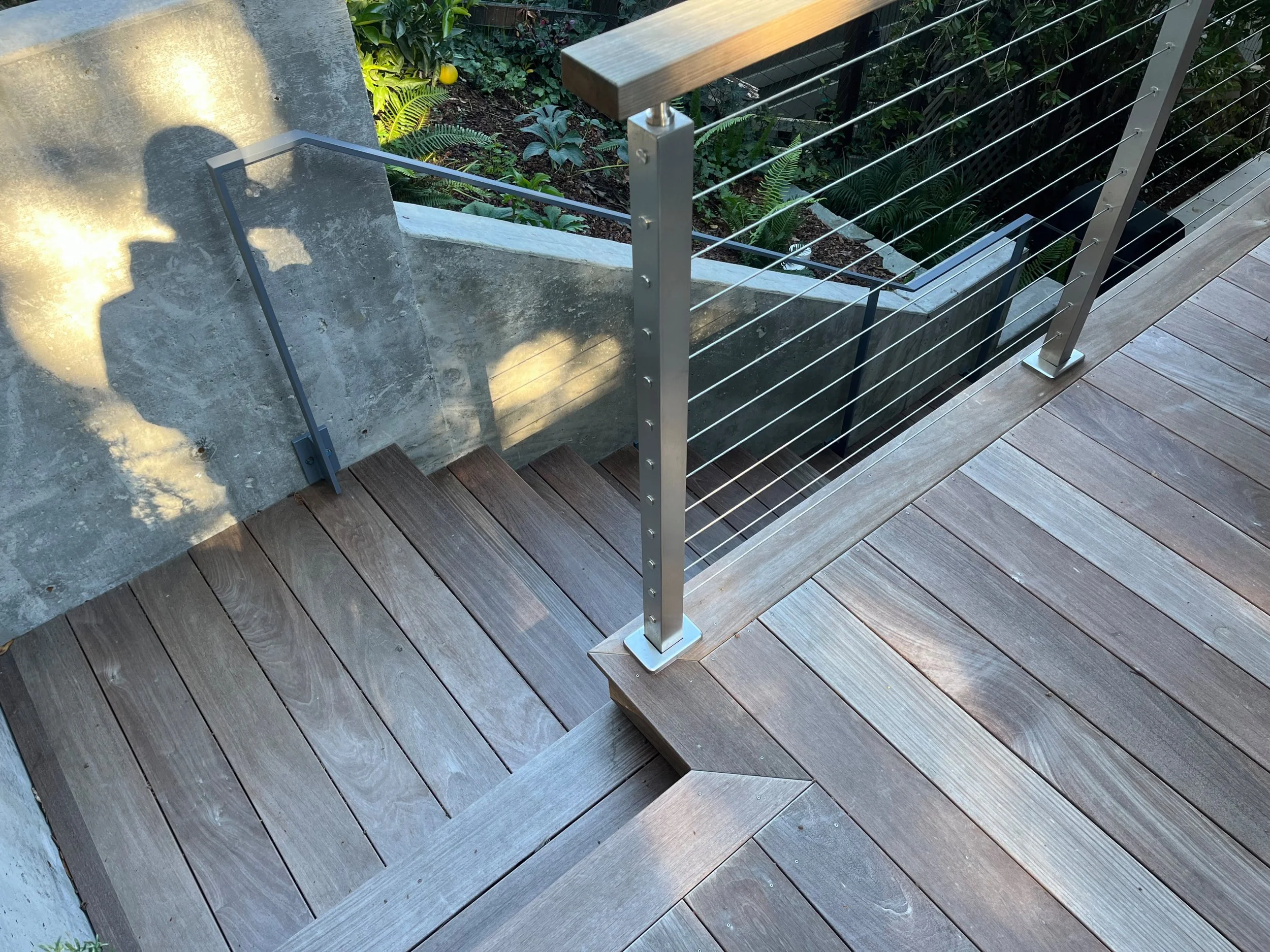 Wooden deck with a metal and cable railing on the edge, overlooking a garden with lush green plants.