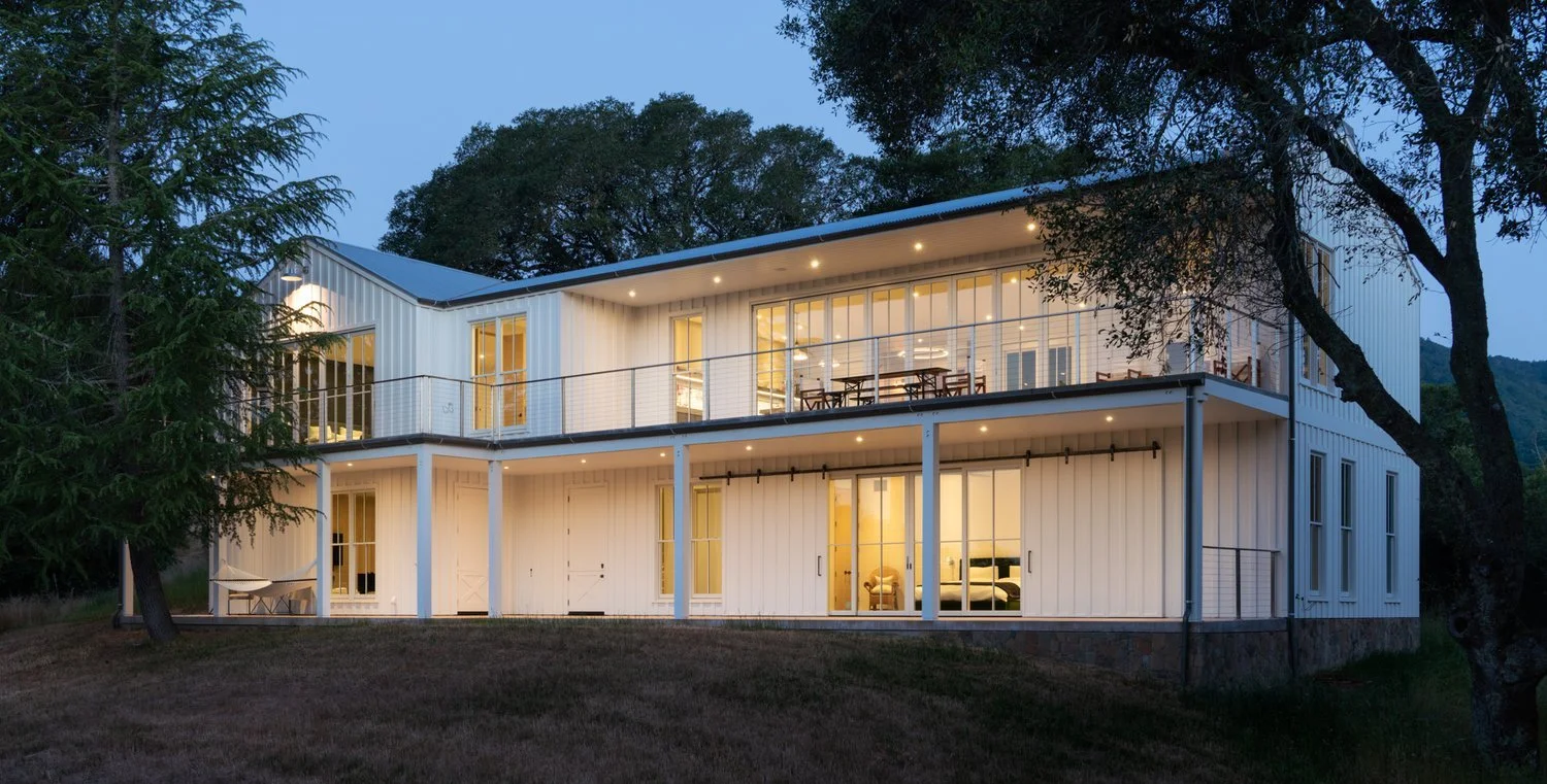 Two-story modern house with large glass windows, white exterior, balcony, and trees surrounding it at dusk.