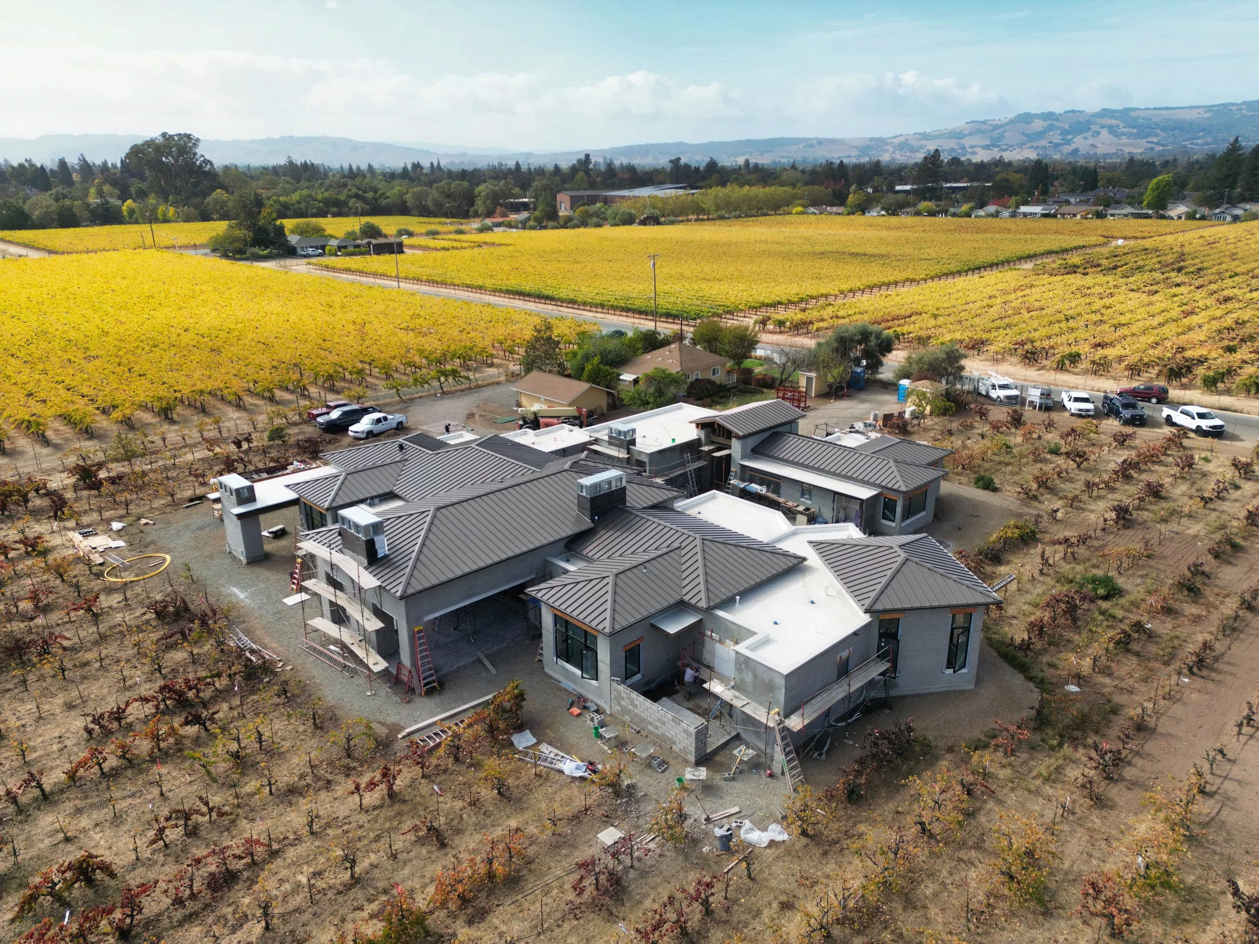 Aerial view of a modern house under construction surrounded by vineyards in a rural area with distant hills and a cloudy sky.