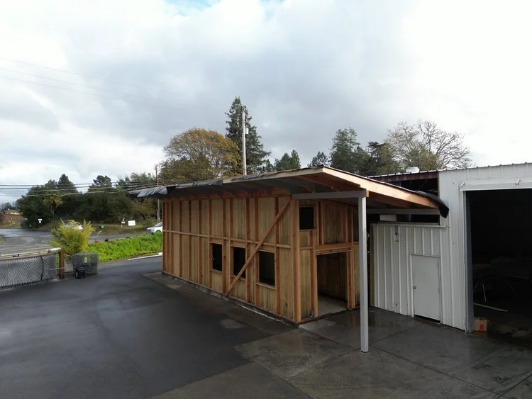 A partially built wooden structure attached to a metal building, with windows and a sloped roof, set on a paved area outdoors.