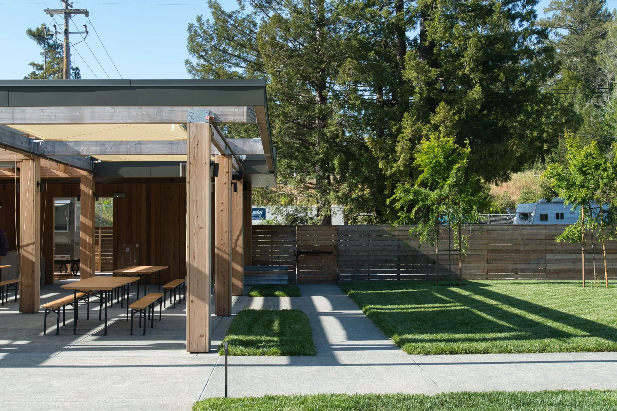 An outdoor patio area with a wooden shelter, tables, and benches, surrounded by a grassy yard and fenced with trees in the background.