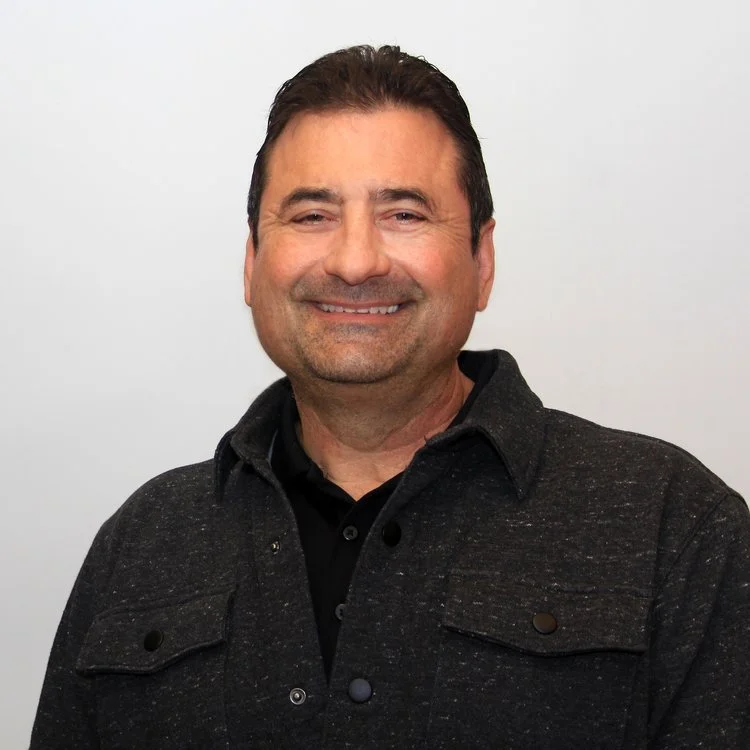 A middle-aged man with brown hair and a beard, smiling, dressed in a black shirt and a dark gray jacket, standing against a plain white background.