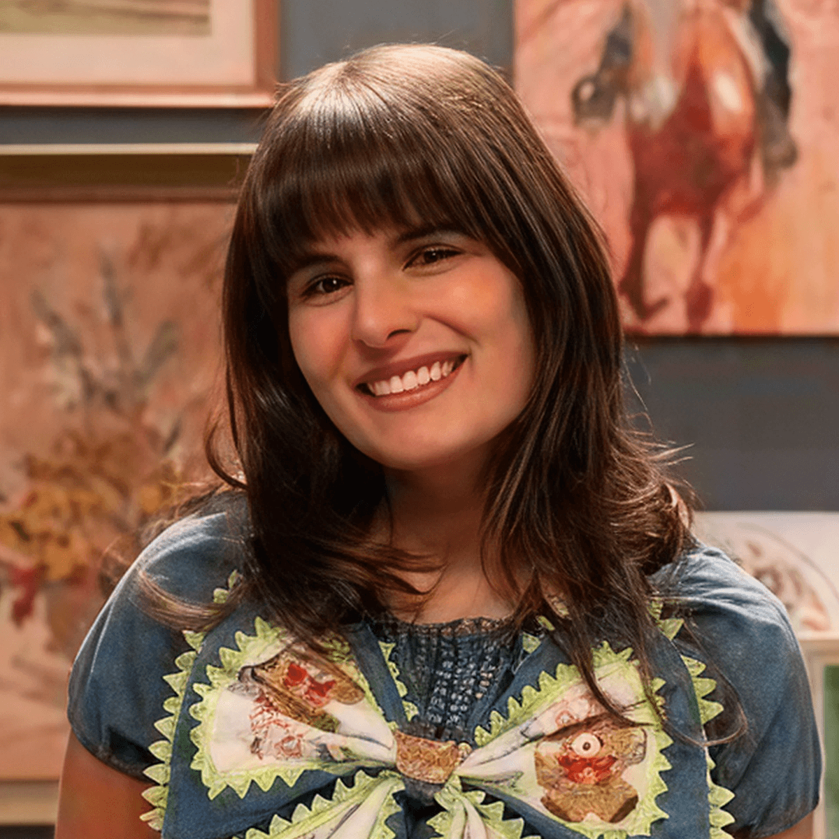 A woman with shoulder-length brown hair, wearing a green collared shirt, standing in front of colorful artwork.