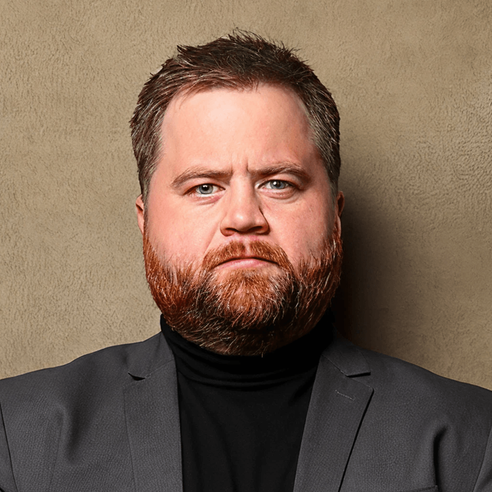 A man with a full beard and mustache, light blue eyes, and light brown hair, wearing a collared shirt with a tiger logo and a navy blazer.
