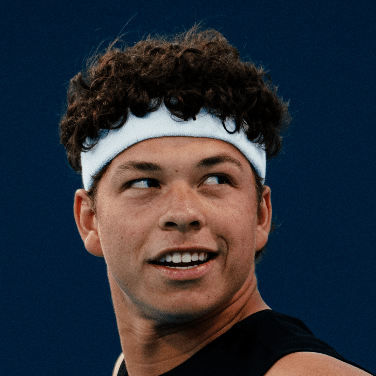 A man with curly hair and a sleeveless white athletic shirt appears to be shouting or calling out during a sports event, with a blurred crowd in the background.