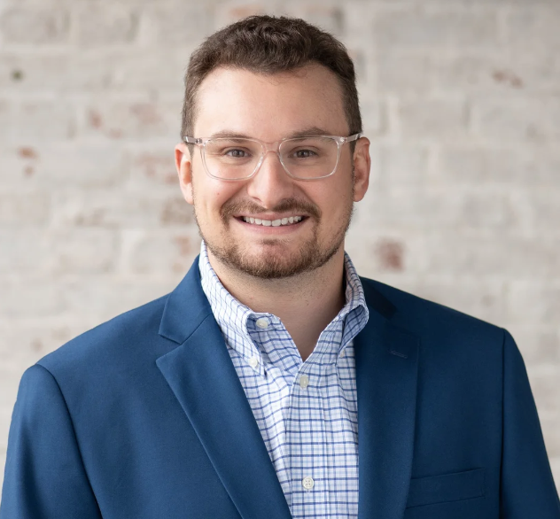 Portrait of a young man with glasses, smiling, in a blue blazer and checked shirt, against a light brick wall background.