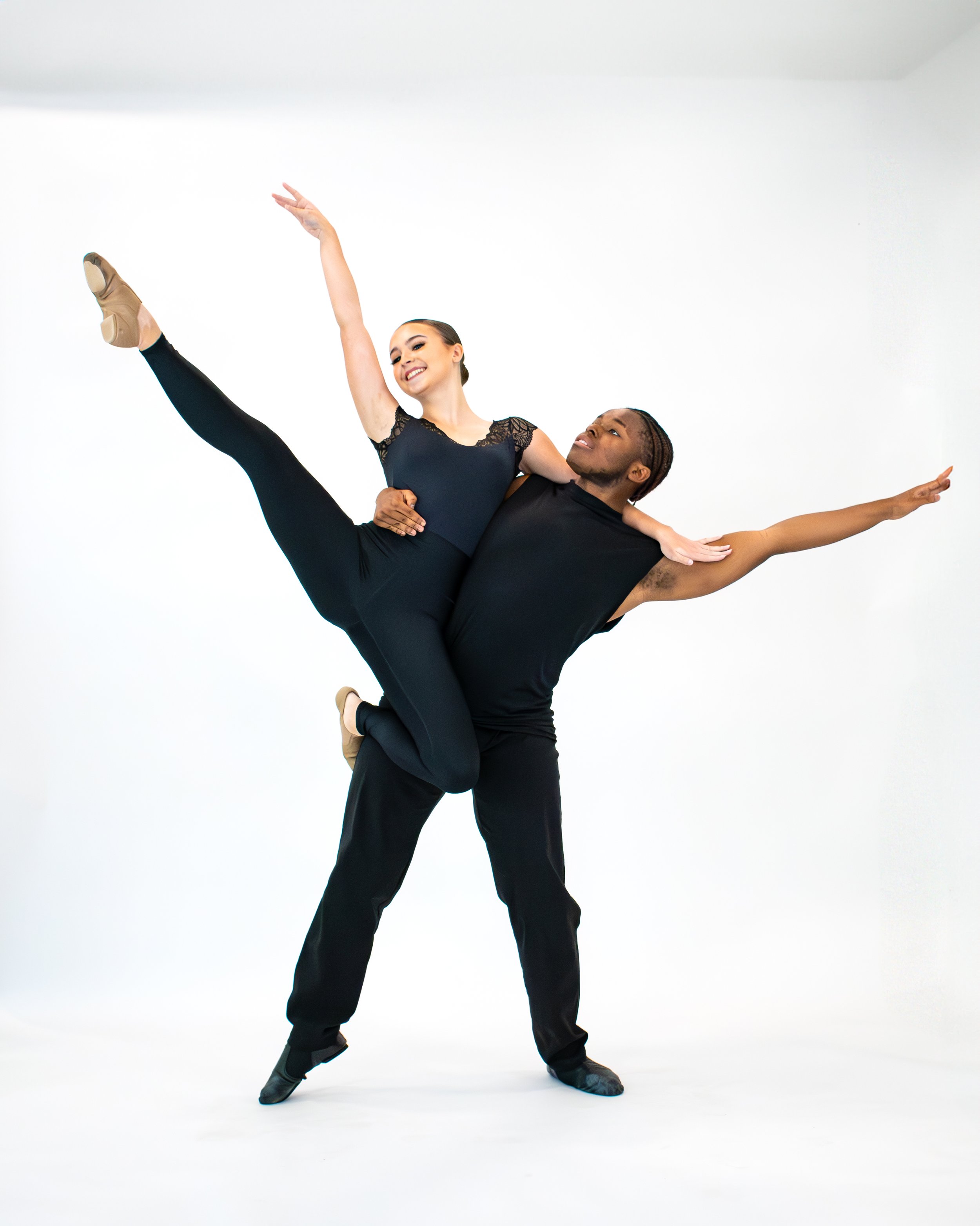 Two ballet dancers performing a lift against a plain white background, one with dark skin and the other with light skin, both wearing black dance attire.
