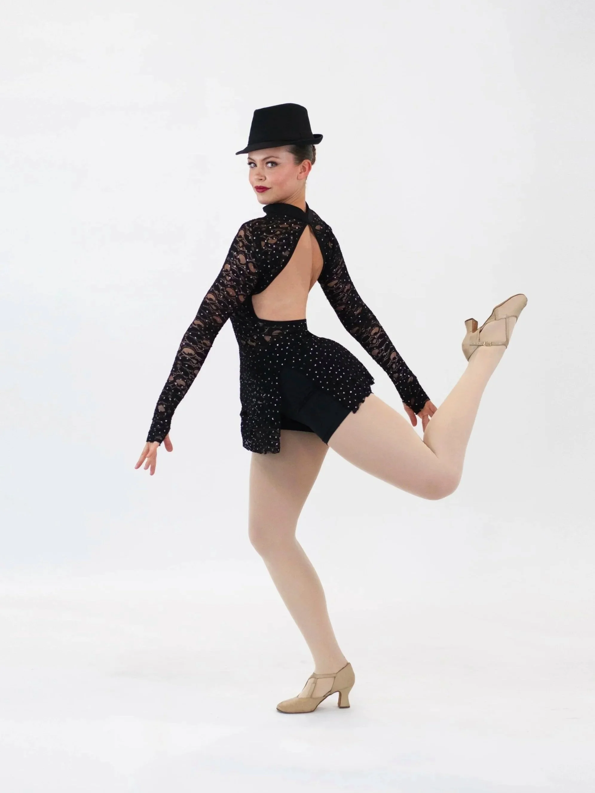 Jazz dancer performing a pose wearing beige character shoes, black dance outfit with a keyhole back, and a black hat, against a white background.