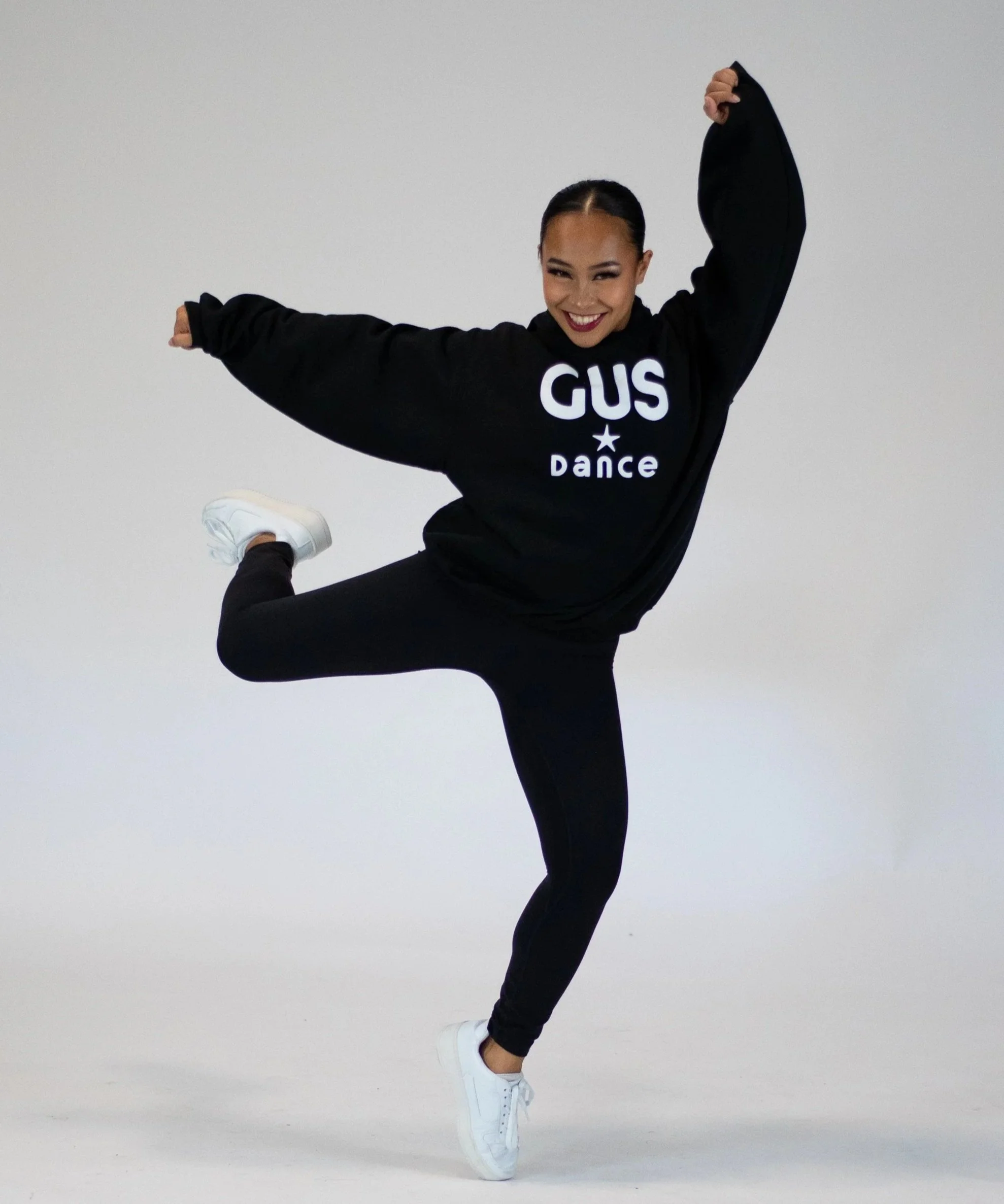A young woman dancing and smiling, wearing a black sweatshirt with 'GUS Dance' written on it, black leggings, and white sneakers, standing against a plain background.