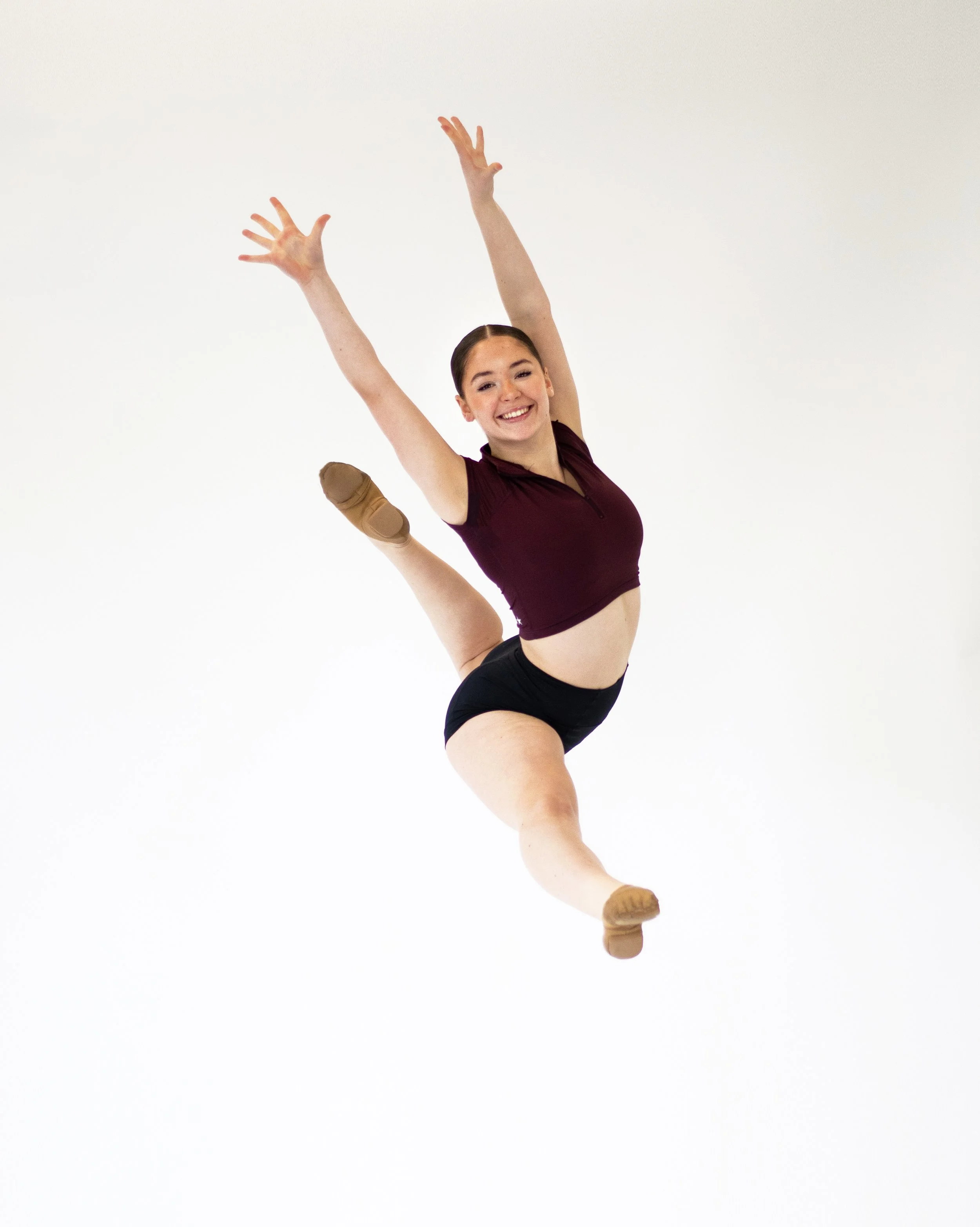 A female dancer in a burgundy top and black shorts is leaping, smiling, against a plain white background.