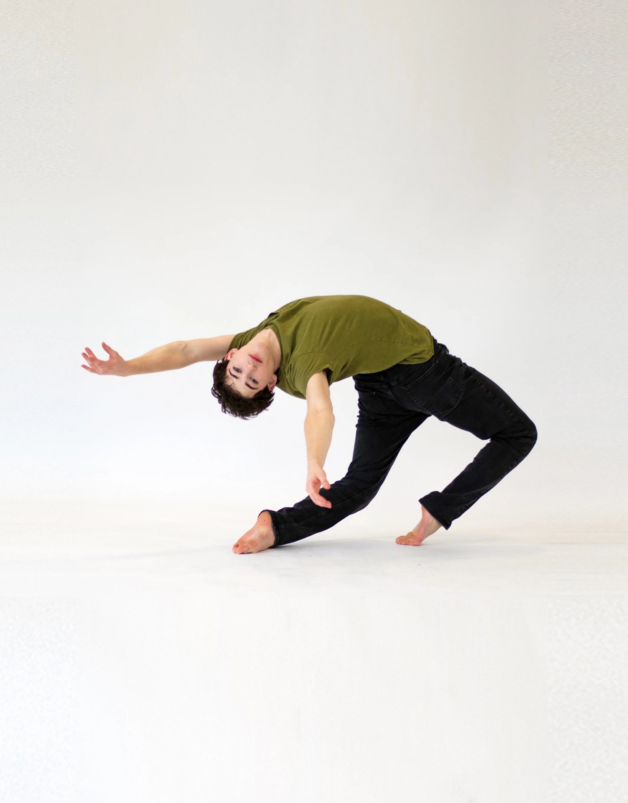 A male dancer leaning sideways with one hand on the floor and the other arm extended upwards, wearing a green shirt and black pants, against a plain white background.