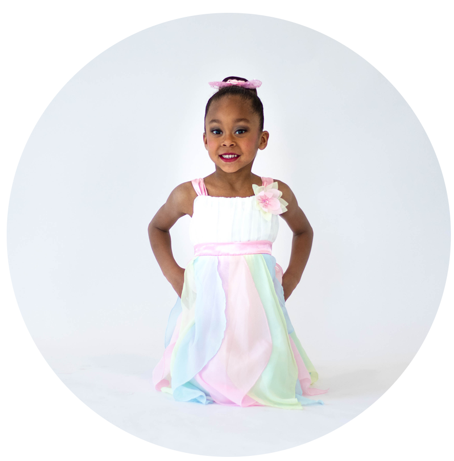 A young girl wearing a pastel rainbow-colored dress with a pink waistband and floral embellishment, smiling against a white background.