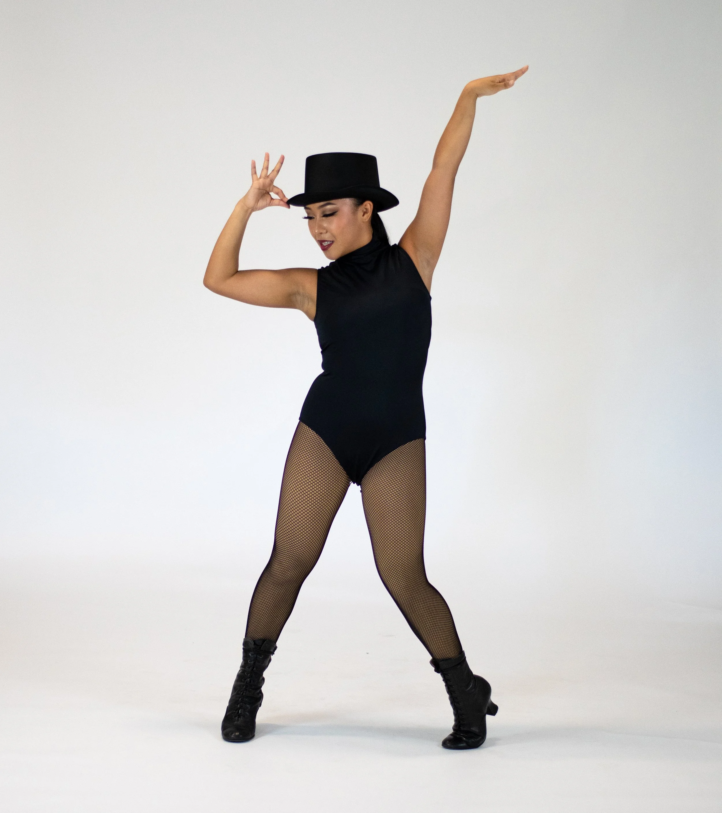 A woman dancing wearing a black top, fishnet tights, black boots, and a black top hat against a plain white background.