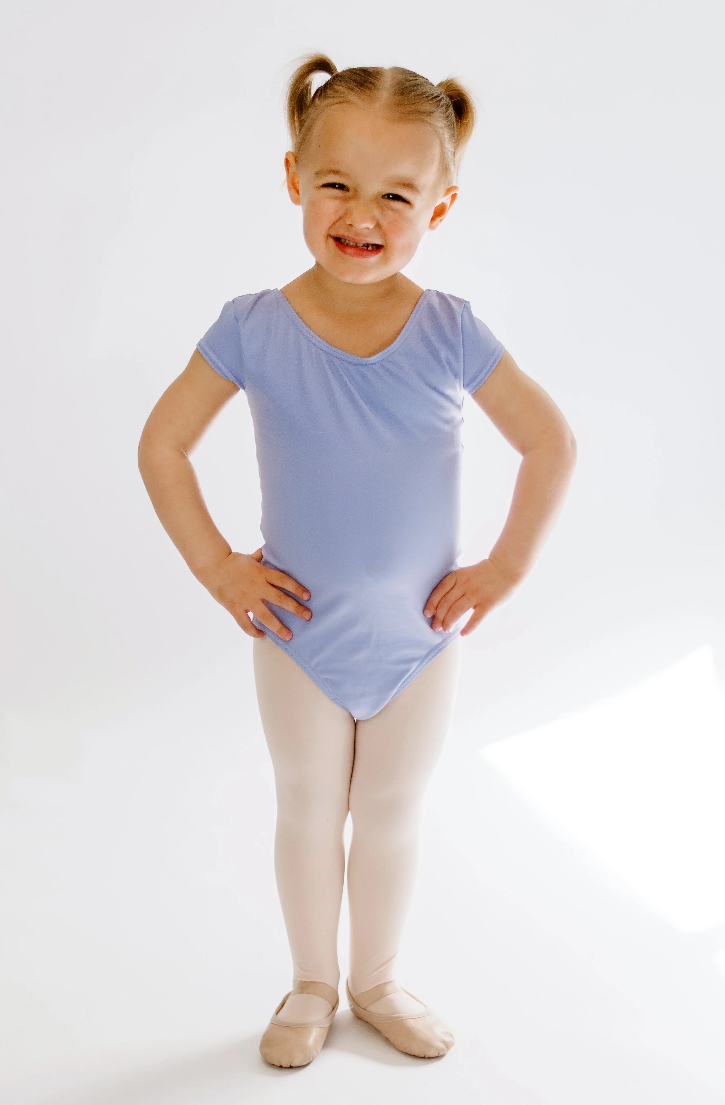 Young girl in ballet attire, standing with hands on hips, smiling, wearing a light blue leotard, ballet slippers, and white tights.