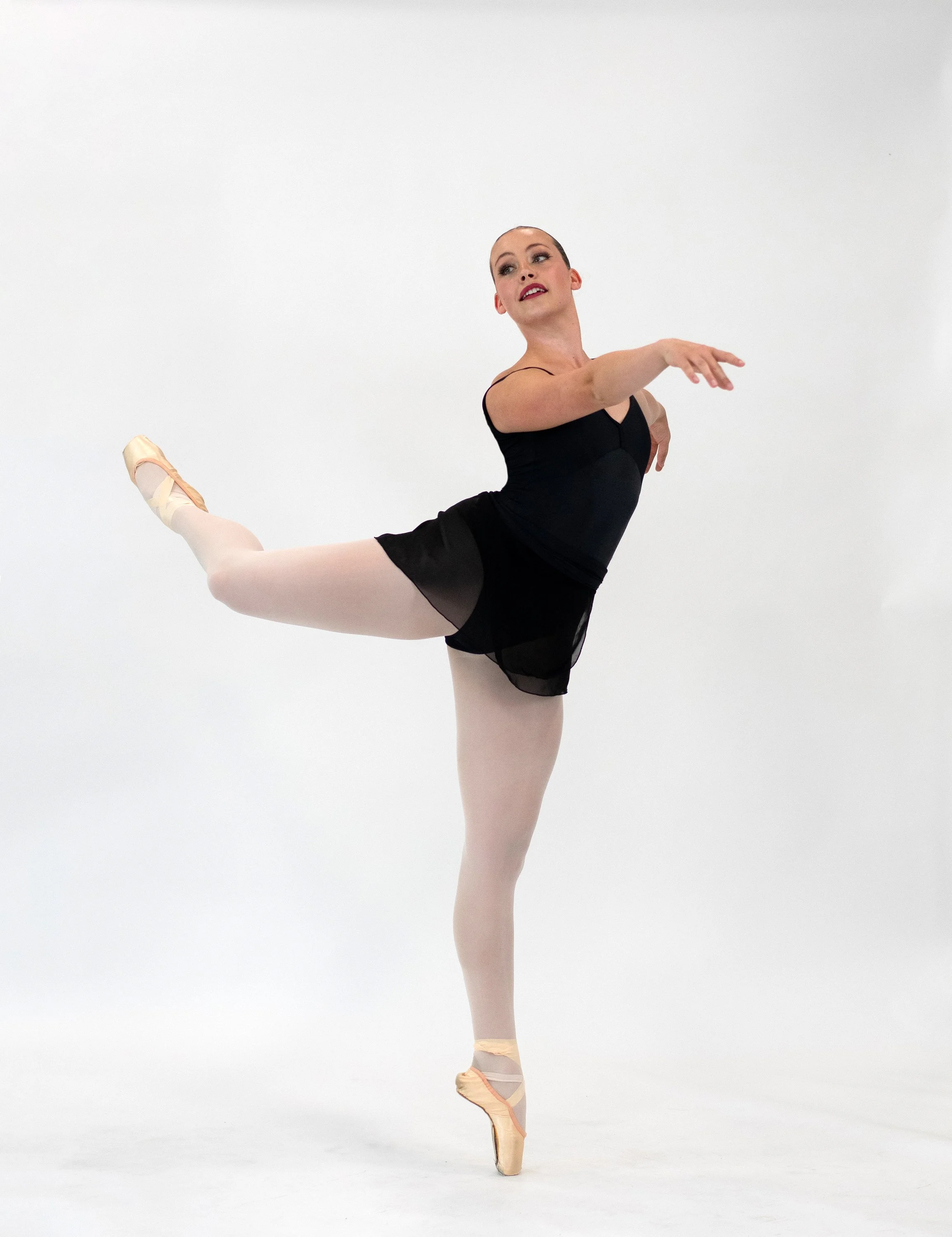 Ballet dancer performing a pose on pointe with one leg lifted and arms extended, wearing a black leotard, pink tights, and pointe shoes, against a plain white background.