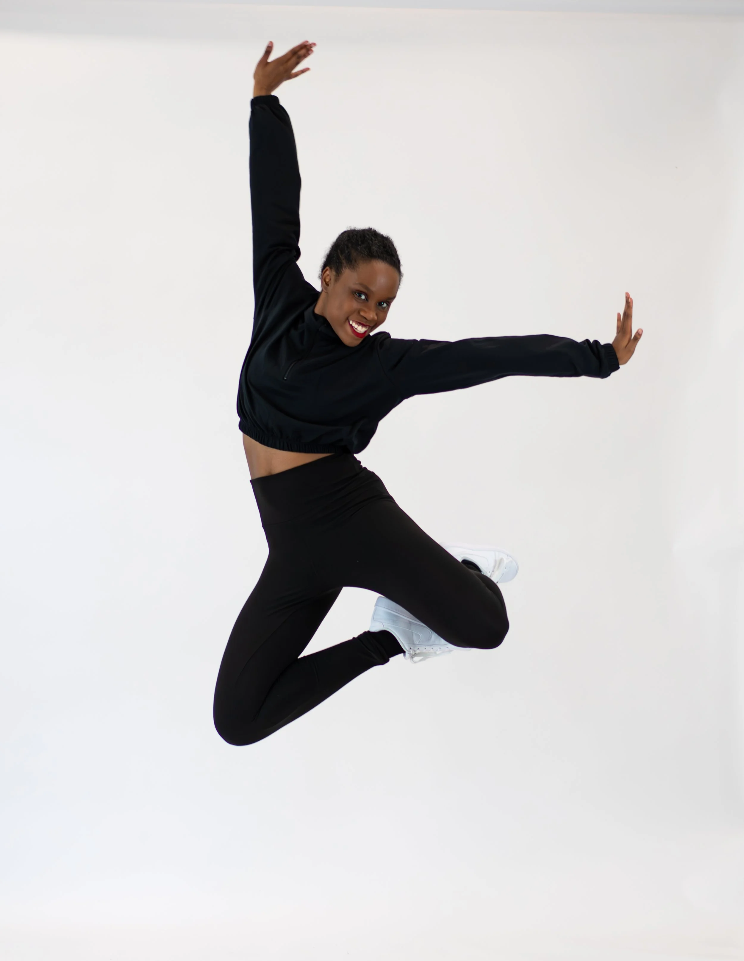 A dancer in black athletic wear performing a pose with one arm raised and the other extended sideways, against a white background.