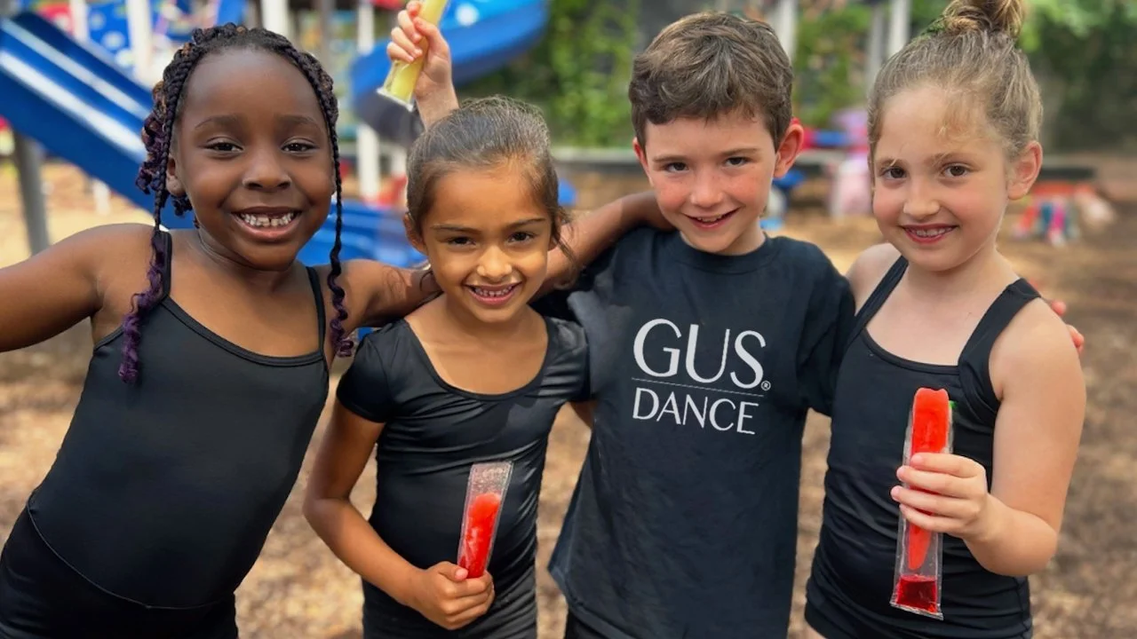 Four children, three girls and one boy, standing together at an outdoor playground, smiling. The girls are holding popsicles, and the boy has his arms around two of them. They are wearing black sports tops, and the background shows a slide and trees.