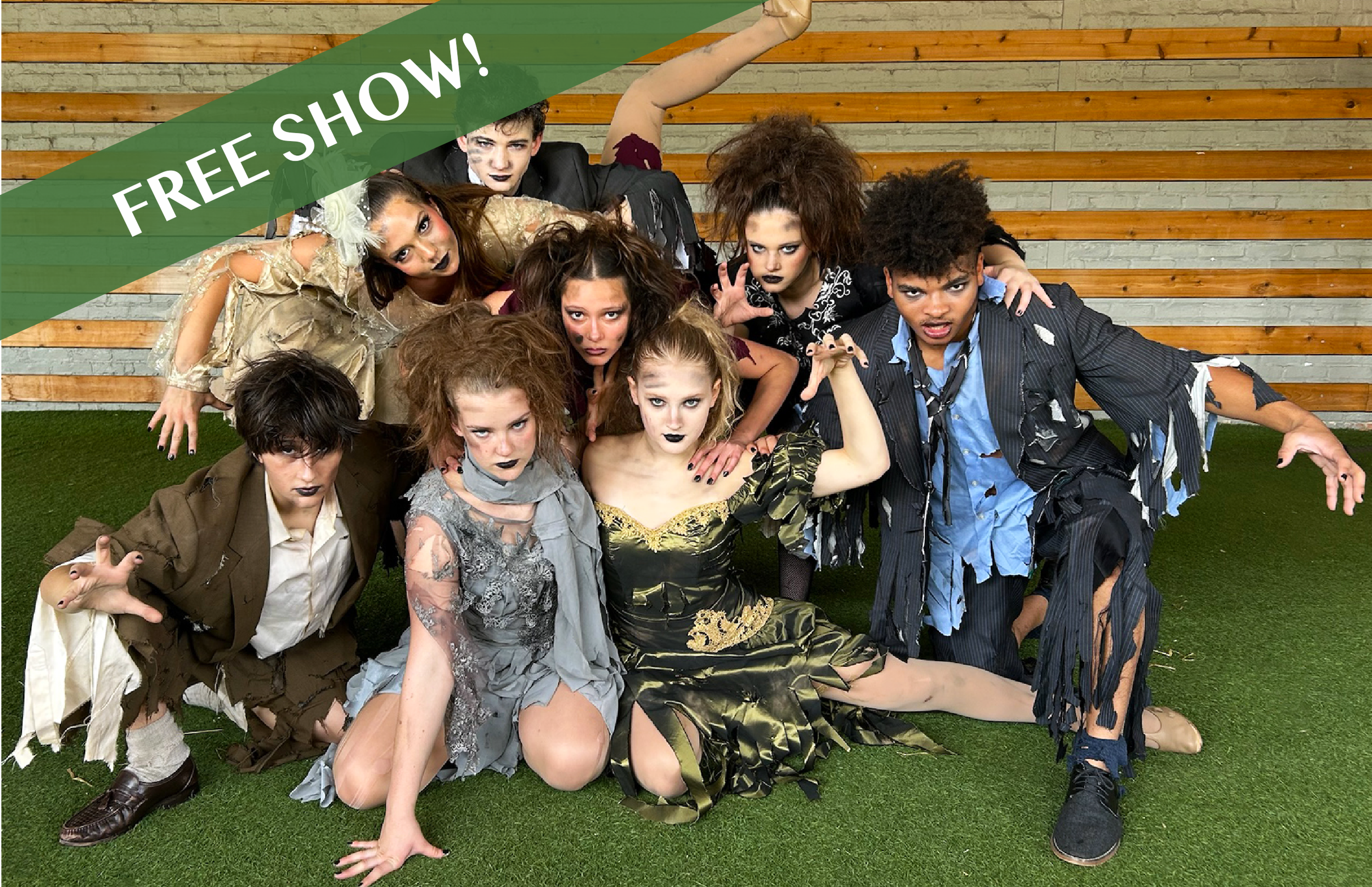 Group of children dressed as zombies in Halloween costumes, crouched on grass, with a wooden wall background.