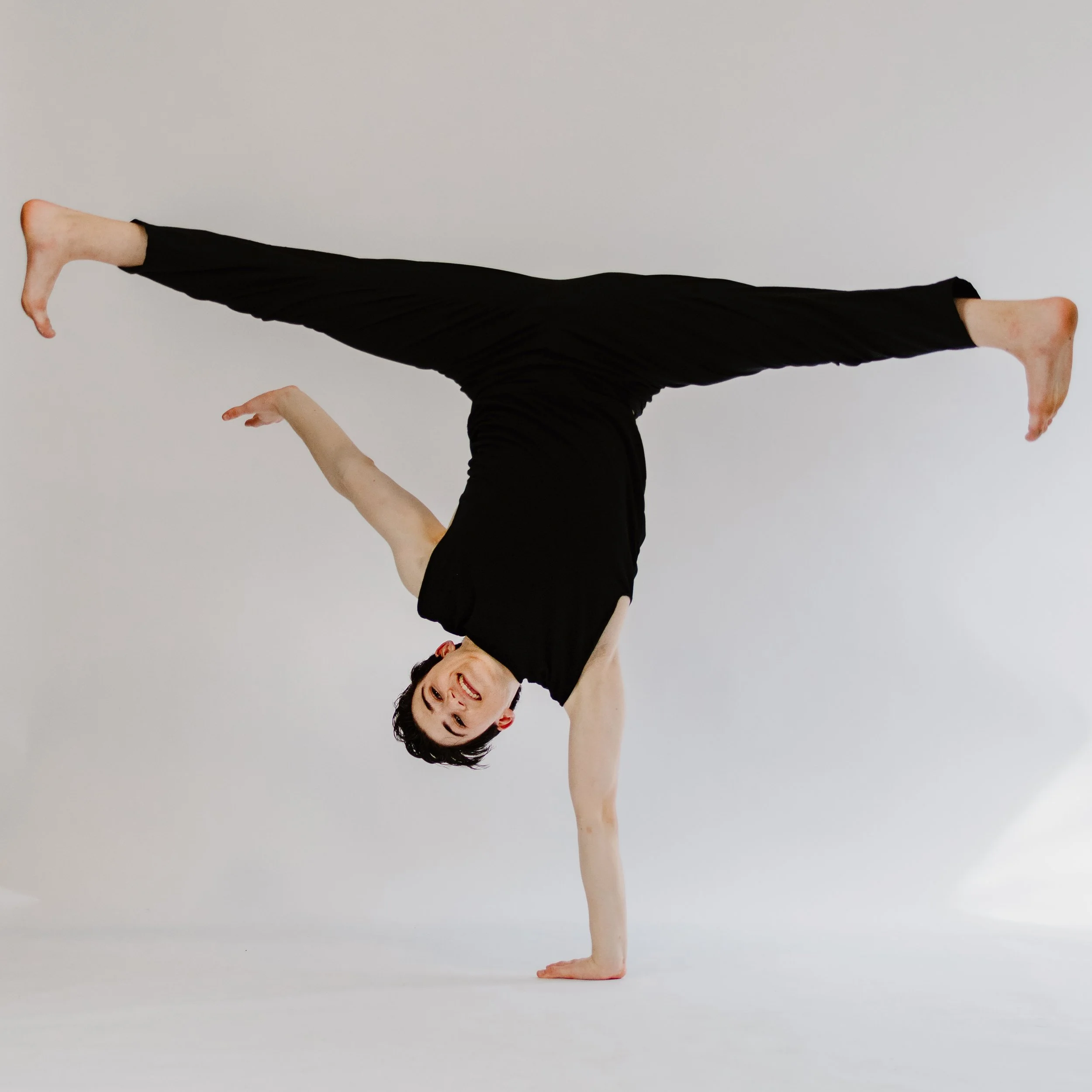 A person performing a yoga handstand against a plain white background, with one hand on the ground and legs extended in a split.