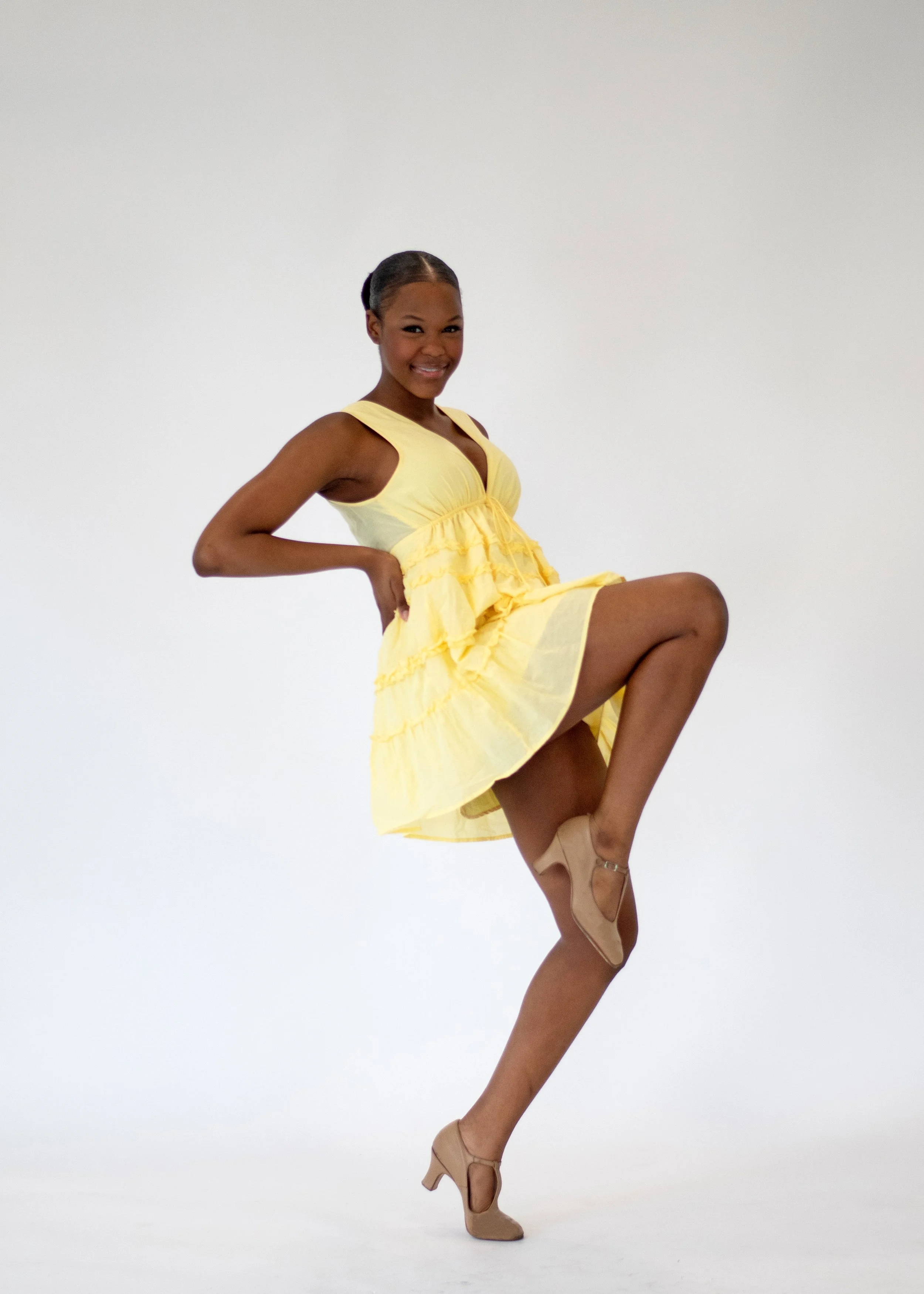 A dancer wearing a yellow dress and beige heels, lifting her right leg with her hand on her hip, smiling against a white background.