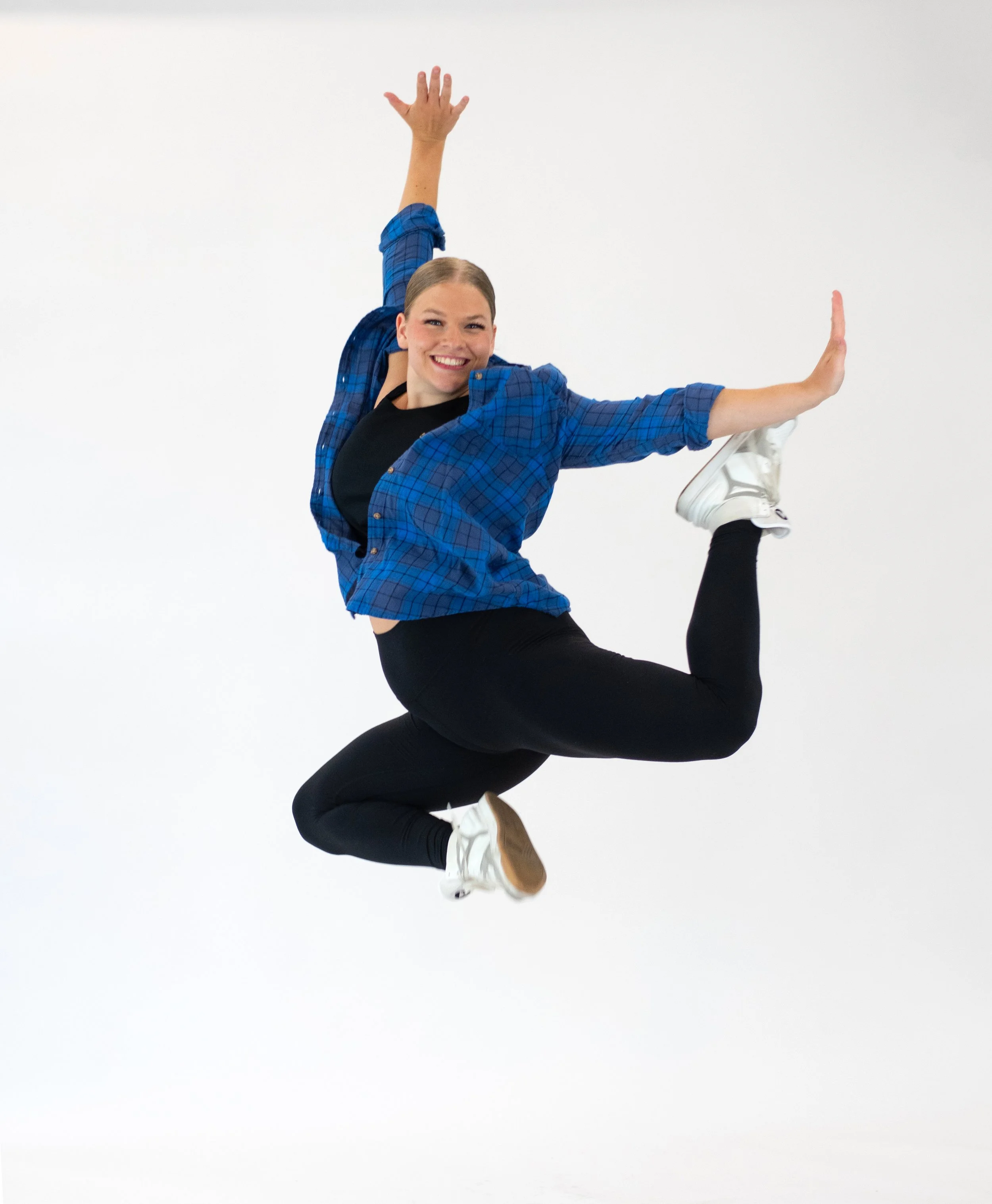 A dancer with hair tied back, wearing a blue plaid shirt, black leggings, and white sneakers, jumping against a plain white background. She is smiling and balancing on one leg while extending her arms and the other leg.