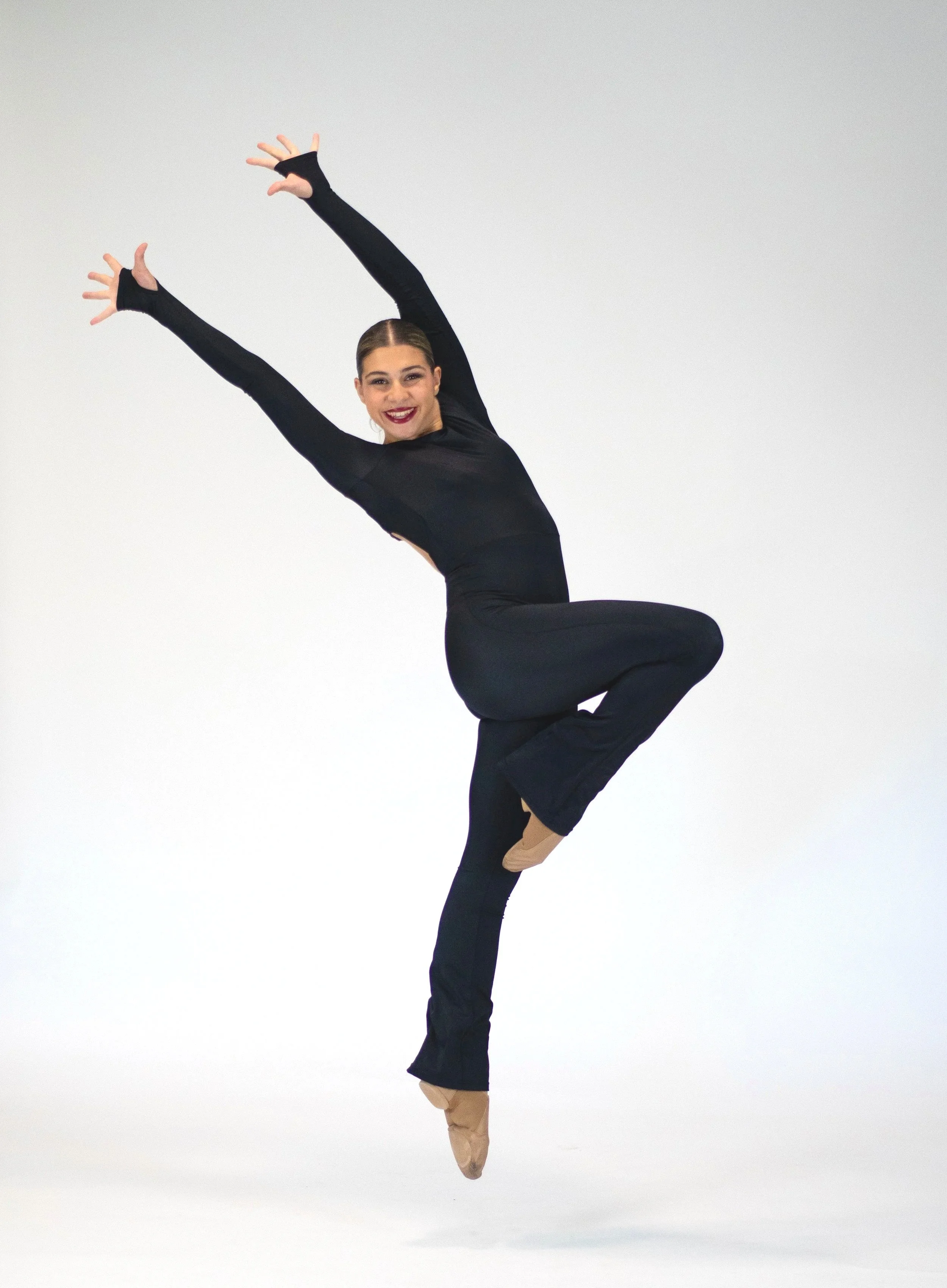 A female dancer balancing on one leg with arms raised, wearing a black outfit with beige jazz shoes, against a plain white background.
