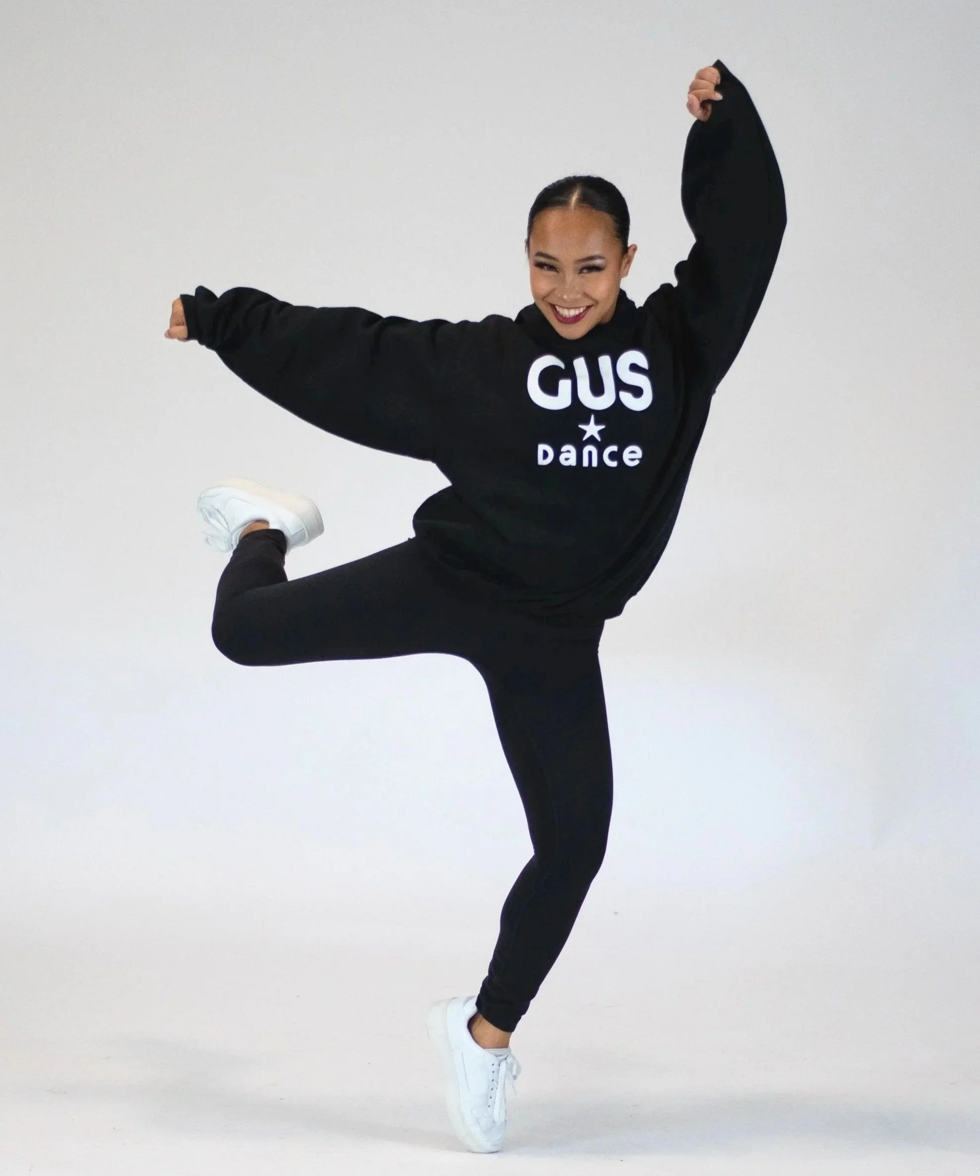 A hip hop dancer in a black hoodie, black pants, and white sneakers against a plain white background, performing a move on one foot with her arms raised and smiling.
